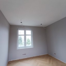 MATS-BUD - Pomalowane na szaro wnętrze pokoju z parkietem w jodełkę, widoczne gniazdka elektryczne i otwory na oświetlenie sufitowe.