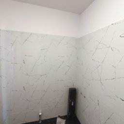 MATS-BUD - Fragment łazienki w Kotlinie z ułożonymi białymi płytkami imitującymi marmur na ścianach oraz czarnymi płytkami na podłodze, widoczne akcesoria glazurnicze i opakowanie fugi.