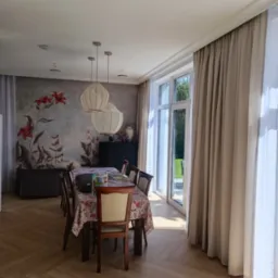 Jasne wnętrze jadalni z parkietem ułożonym w jodełkę, kwiatowym motywem na ścianie, trzema lampami wiszącymi nad stołem przykrytym kwiecistym obrusem i zasłonami w oknach.