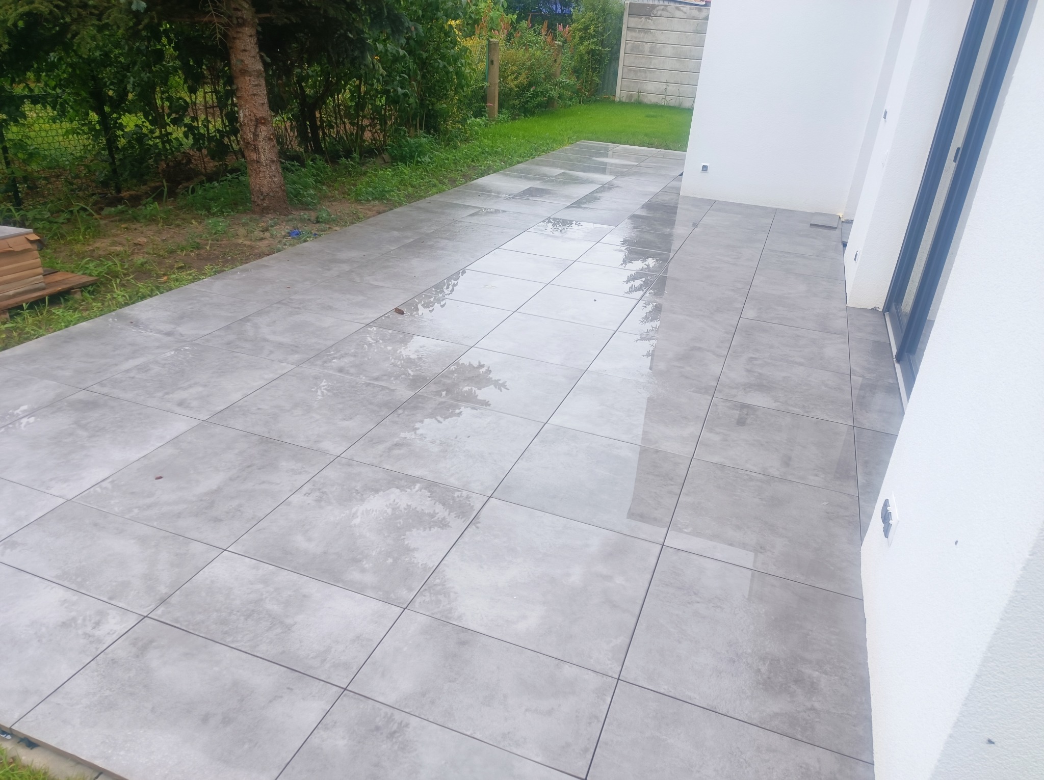 Nowoczesny taras z szarych, kwadratowych płytek, częściowo mokrych po deszczu, przylegający do białej ściany budynku i zielonego trawnika.