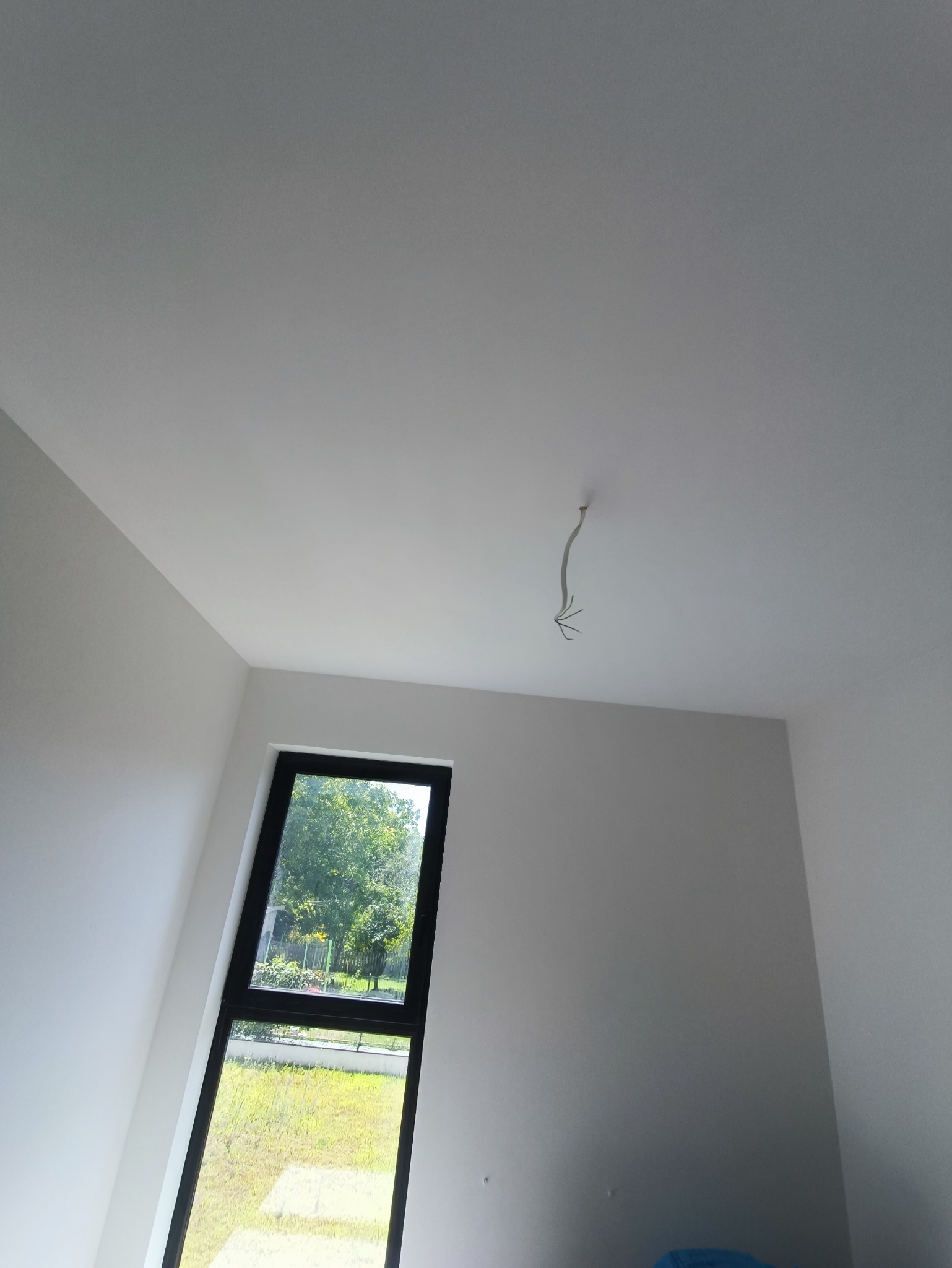 Wnętrze pokoju z białymi ścianami i sufitem, czarną ramą okienną z widokiem na zieleń, oraz wystającymi kablami z sufitu.