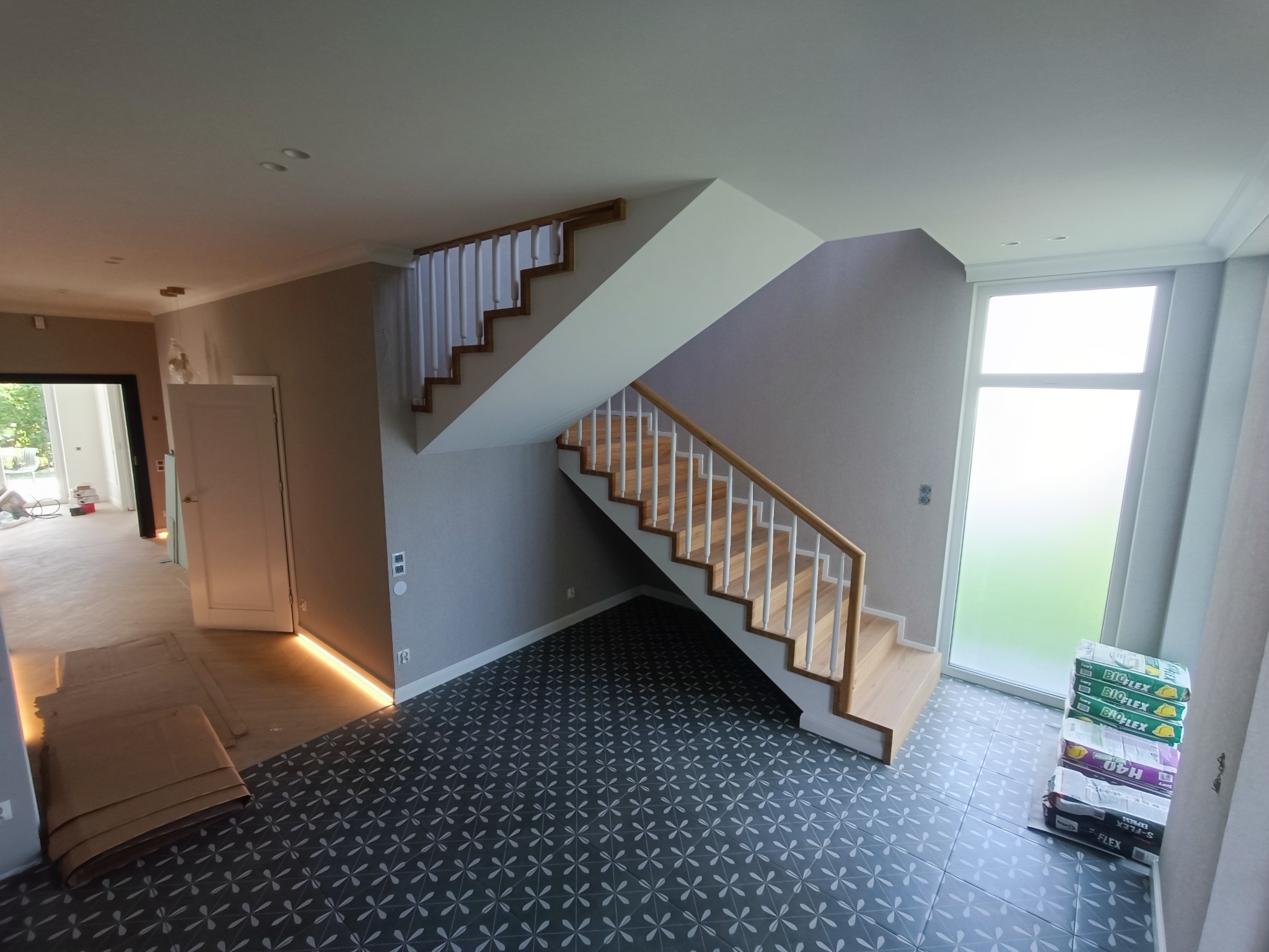 Nowoczesne wnętrze po remoncie: szare płytki podłogowe we wzory, drewniane schody z białymi balustradami, listwy przypodłogowe z oświetleniem LED, paczki kleju do płytek przy oknie.