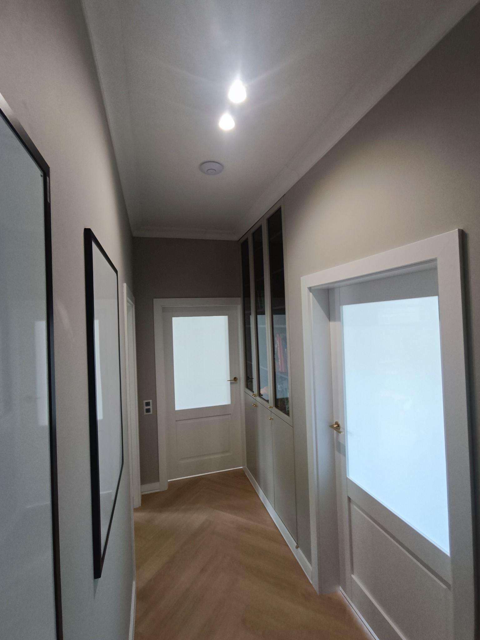 Korytarz z parkietem ułożonym w jodełkę, zabudowaną szafą wnękową z przeszklonymi drzwiami, białymi drzwiami wewnętrznymi z szybami i minimalistycznymi obrazami w czarnych ramach na ścianach.