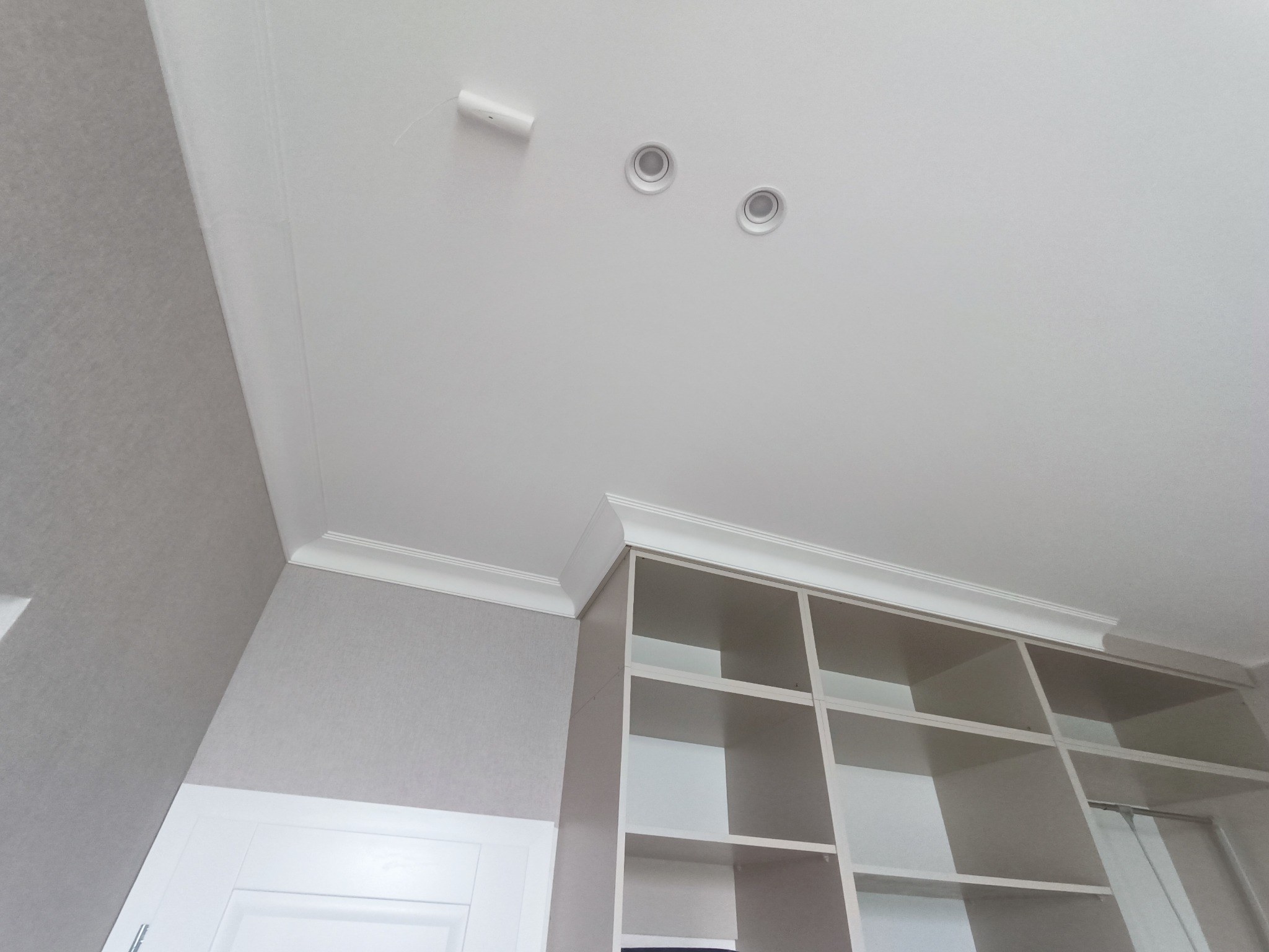 Wnętrze pomieszczenia z białym sufitem, ozdobnym sztukateryjnym gzymsem, wbudowanym regałem z półkami i punktowym oświetleniem.