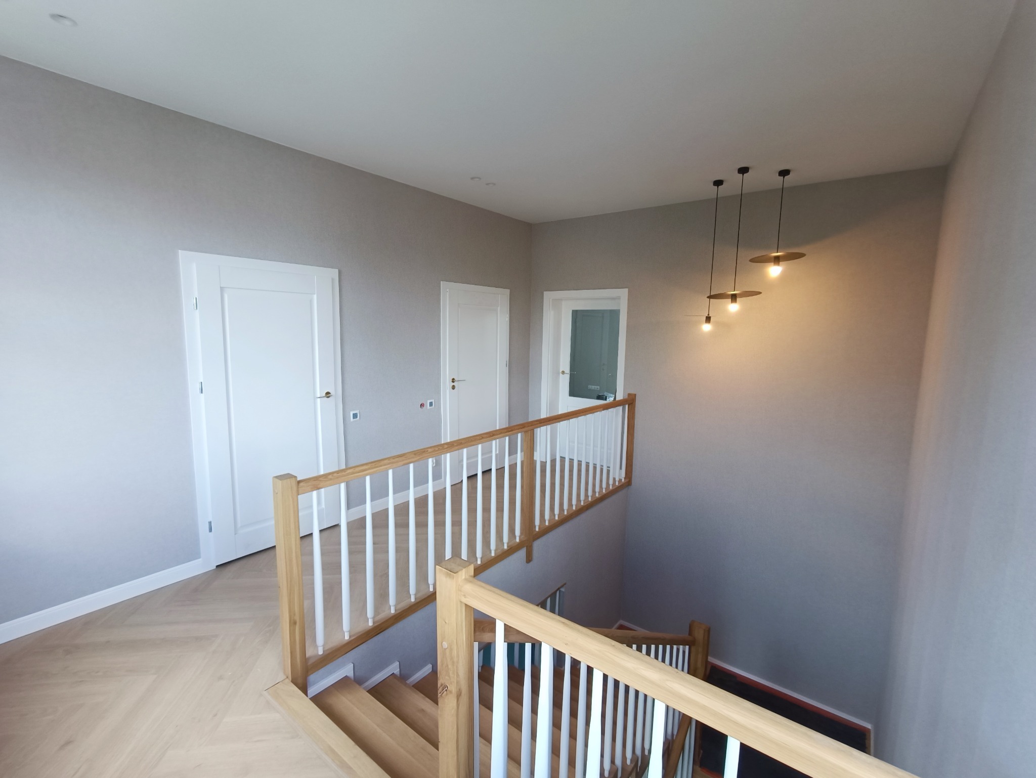 Jasne wnętrze domu z drewnianymi schodami, białymi drzwiami i szarą ścianą, oświetlone trzema nowoczesnymi lampami wiszącymi.