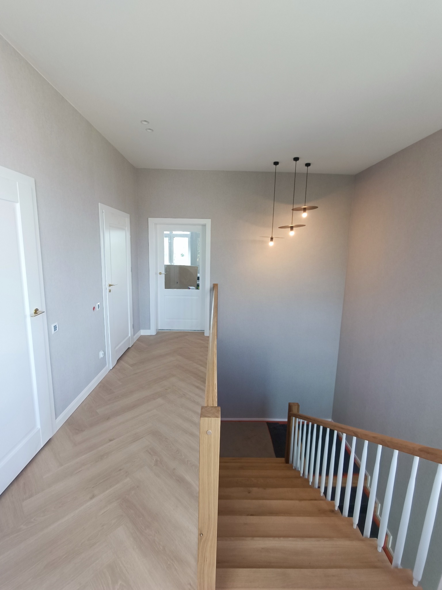 Jasne wnętrze domu z drewnianymi schodami z białymi tralkami, parkietem ułożonym w jodełkę, białymi drzwiami i minimalistycznymi lampami wiszącymi.