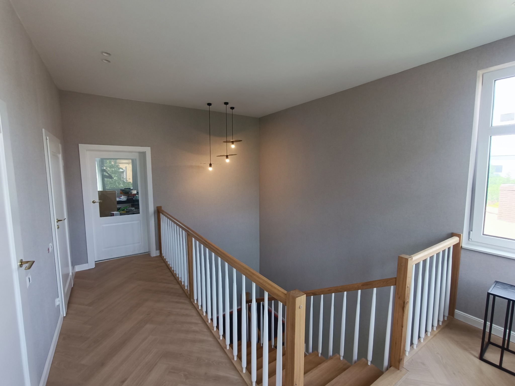 Korytarz z drewnianą podłogą ułożoną w jodełkę, szare ściany i białe drzwi, widoczne schody z drewnianą balustradą i białymi tralkami oraz nowoczesne lampy wiszące.