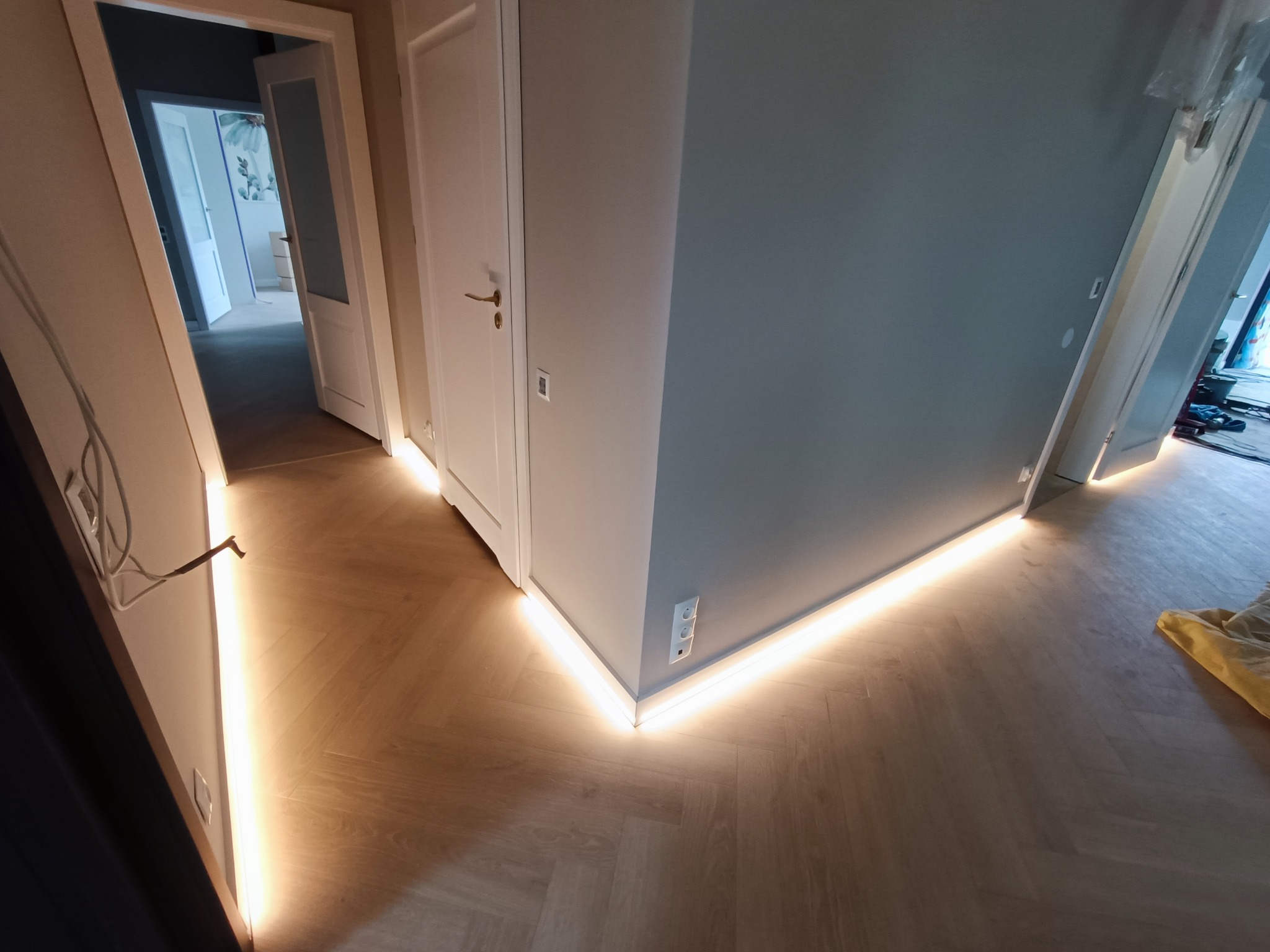Korytarz z podświetleniem LED wzdłuż podłogi, parkiet ułożony w jodełkę, szare ściany, białe drzwi z widocznymi gniazdkami elektrycznymi.