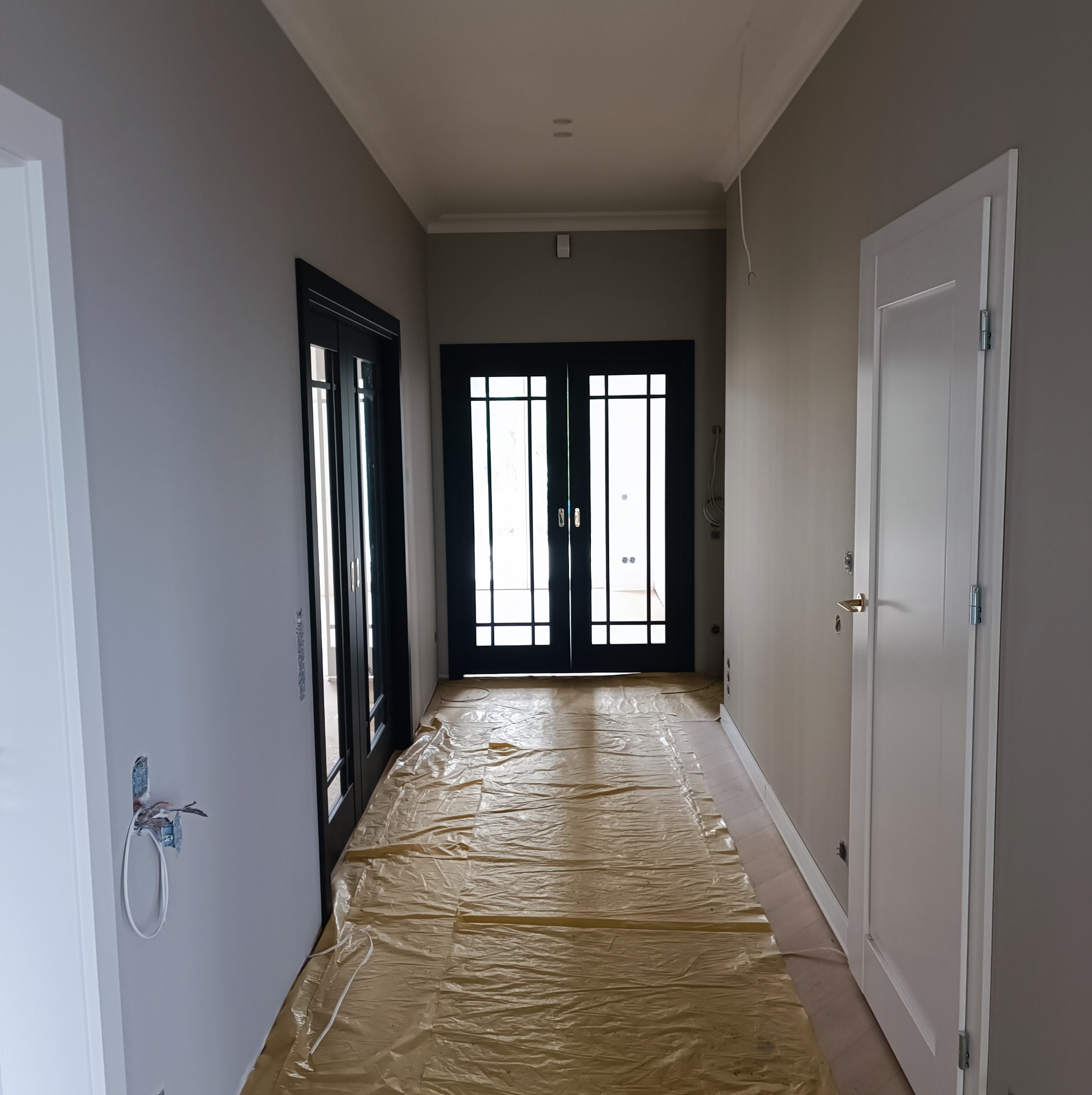 Korytarz w trakcie malowania ścian na szaro, podłoga zabezpieczona folią, widoczne czarne przeszklone drzwi i białe drzwi po prawej stronie, wystające kable ze ściany po lewej.