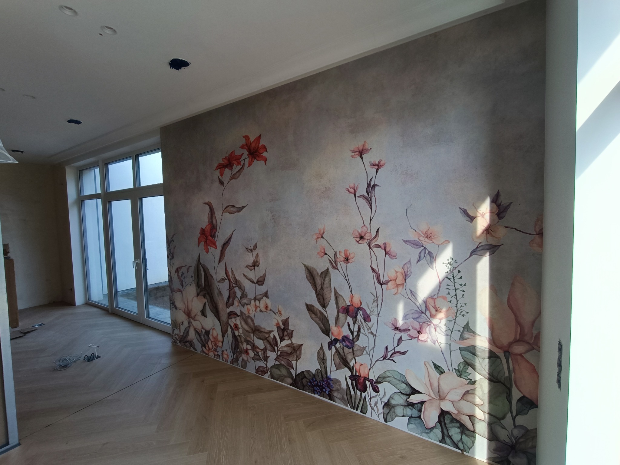 Ściana z dekoracyjną tapetą w kwiaty w nowym wnętrzu z parkietem ułożonym w jodełkę francuską, widoczne przygotowane otwory na oświetlenie.