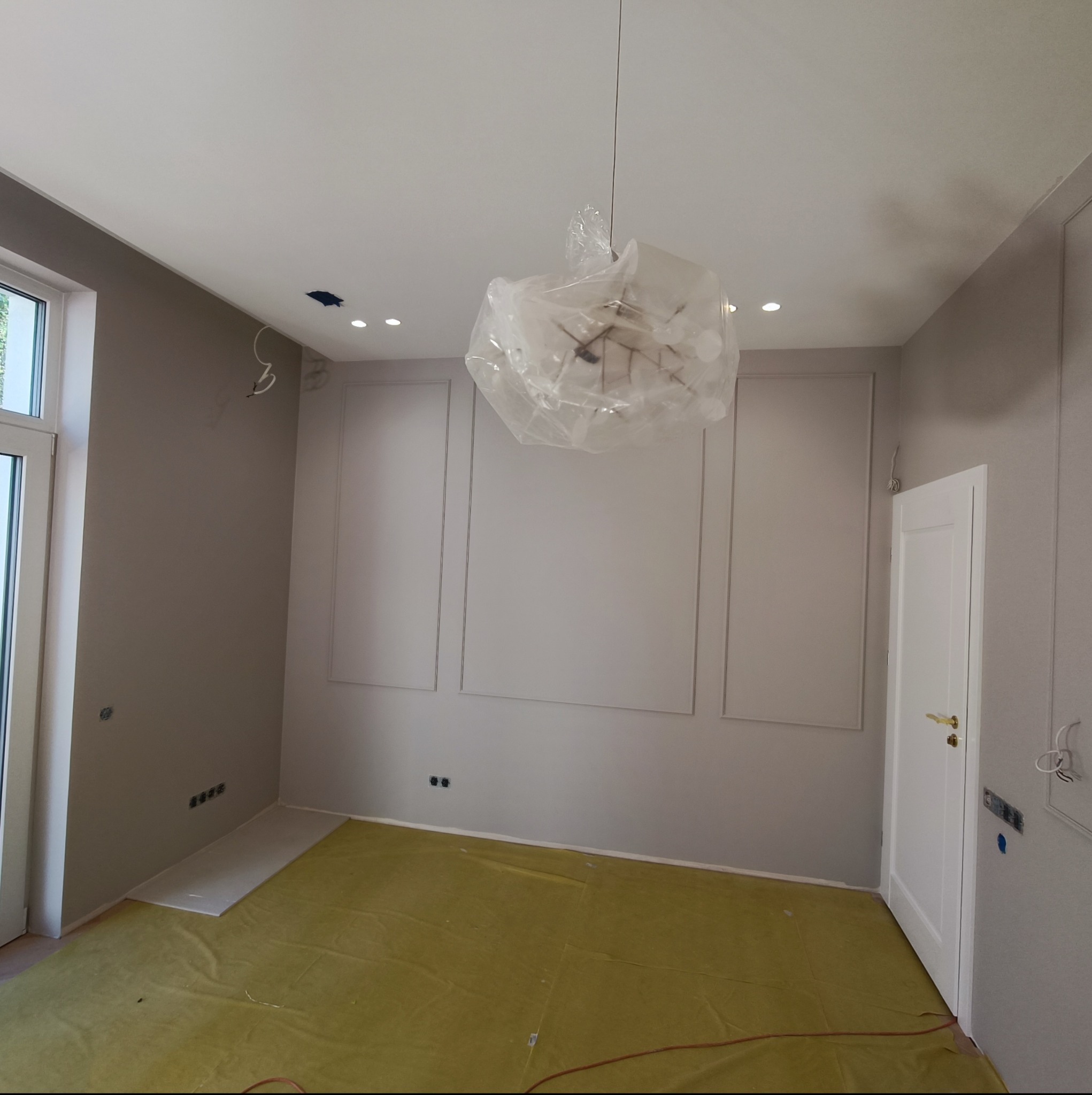 Pokój w trakcie prac wykończeniowych: ściany pomalowane na szaro, dekoracyjne listwy ścienne, podłoga zabezpieczona folią, lampa wisząca owinięta folią ochronną, widoczne puszki elektryczne...