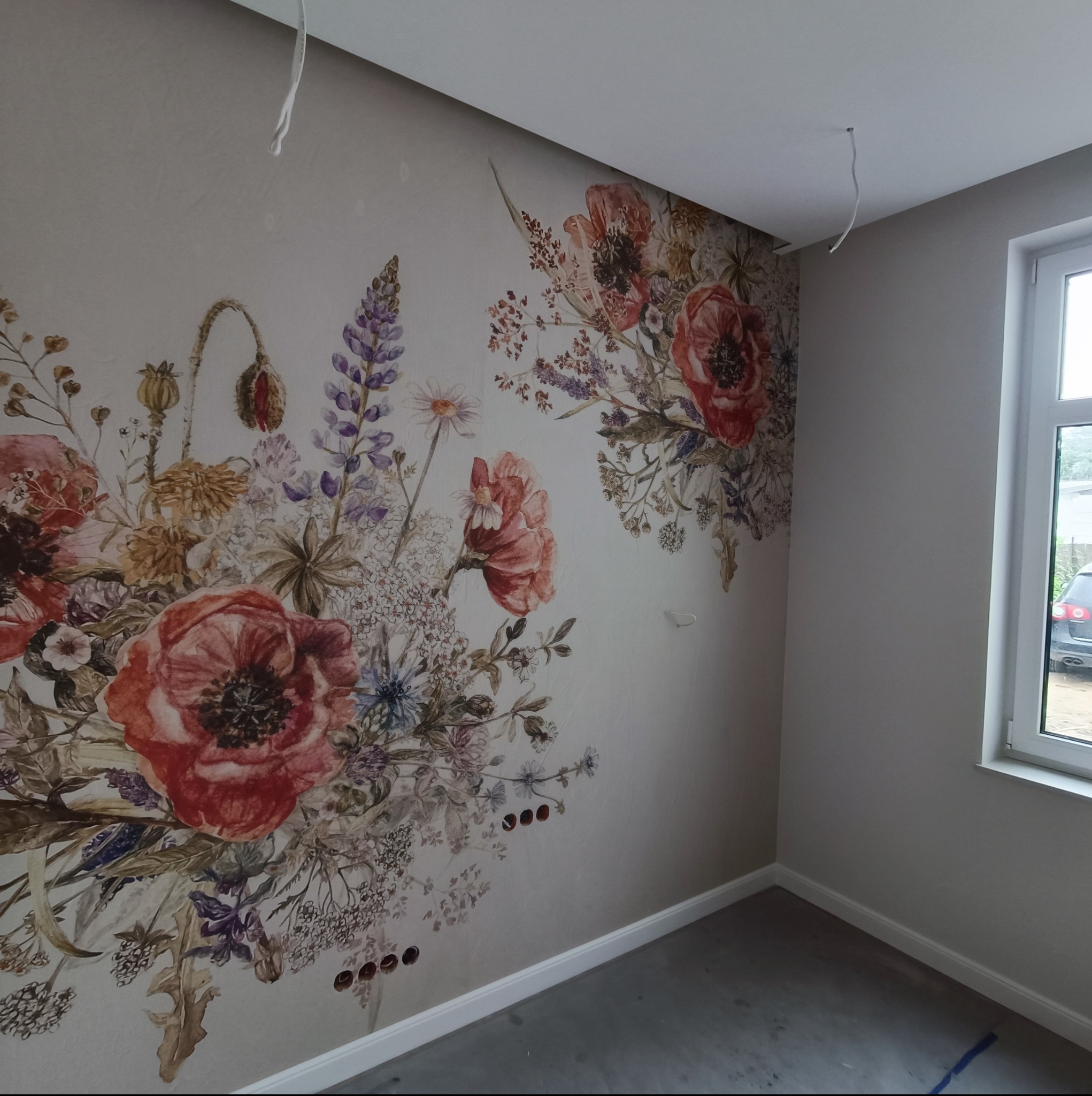 Ściana w pokoju z dekoracyjną tapetą w kwiatowe motywy, widoczne gniazdka elektryczne i wystające przewody z sufitu, okno w tle.