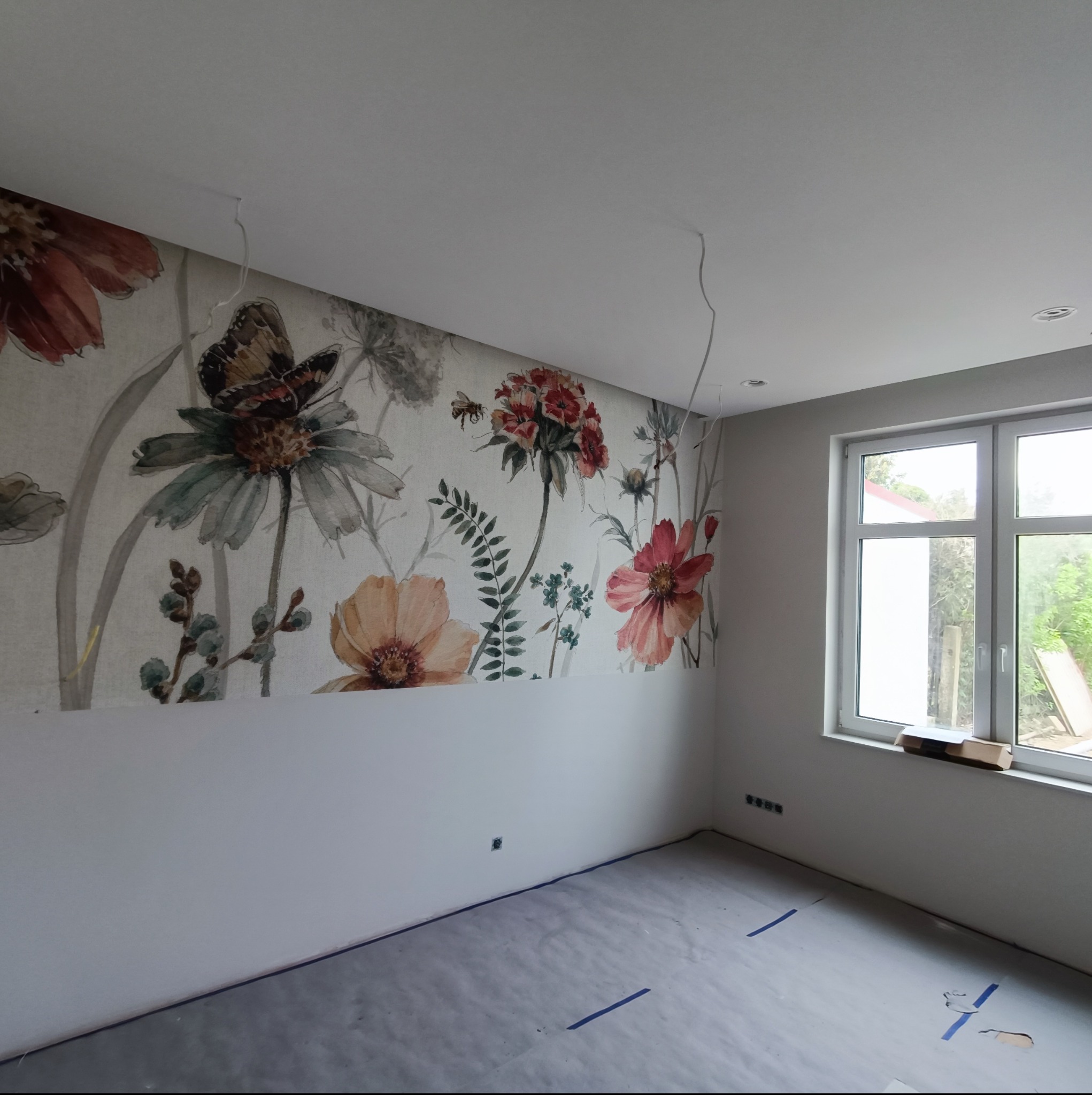 Pokój w trakcie prac, z częściowo położoną tapetą w kwiaty i motyle na górnej części ściany, zabezpieczona podłoga folią malarską, widoczne kable zwisające z sufitu, okno z kartonem na parapecie.