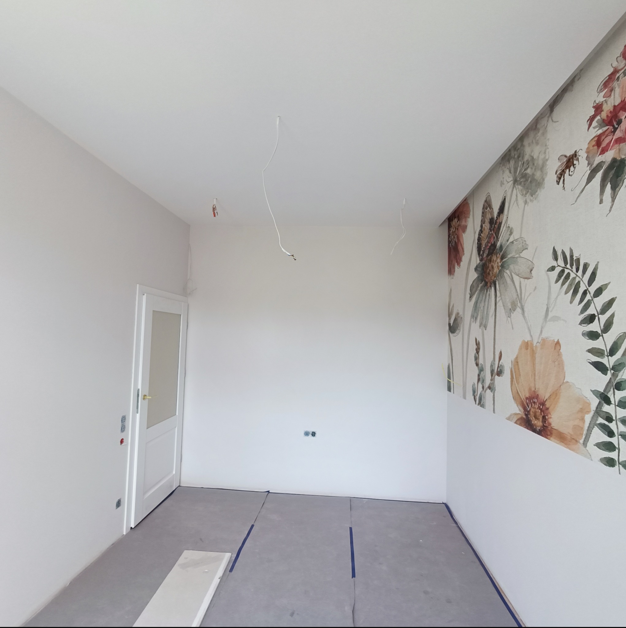 Wnętrze pokoju w Kotlin w trakcie wykańczania: ściany pomalowane na biało, na jednej ścianie tapeta w kwiaty, zabezpieczona folią podłoga, wystające kable z sufitu.