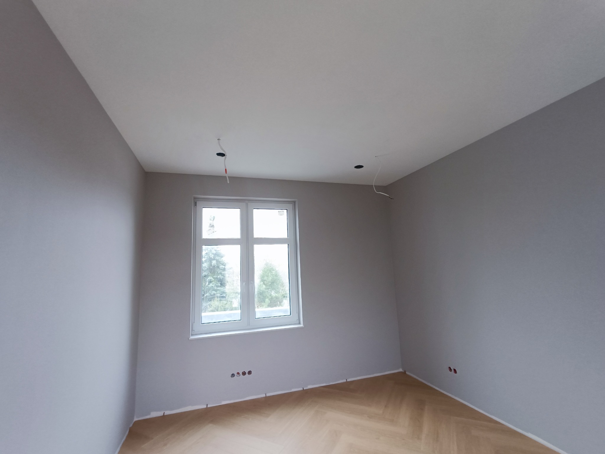 Pomalowane na szaro wnętrze pokoju z parkietem w jodełkę, widoczne gniazdka elektryczne i otwory na oświetlenie sufitowe.