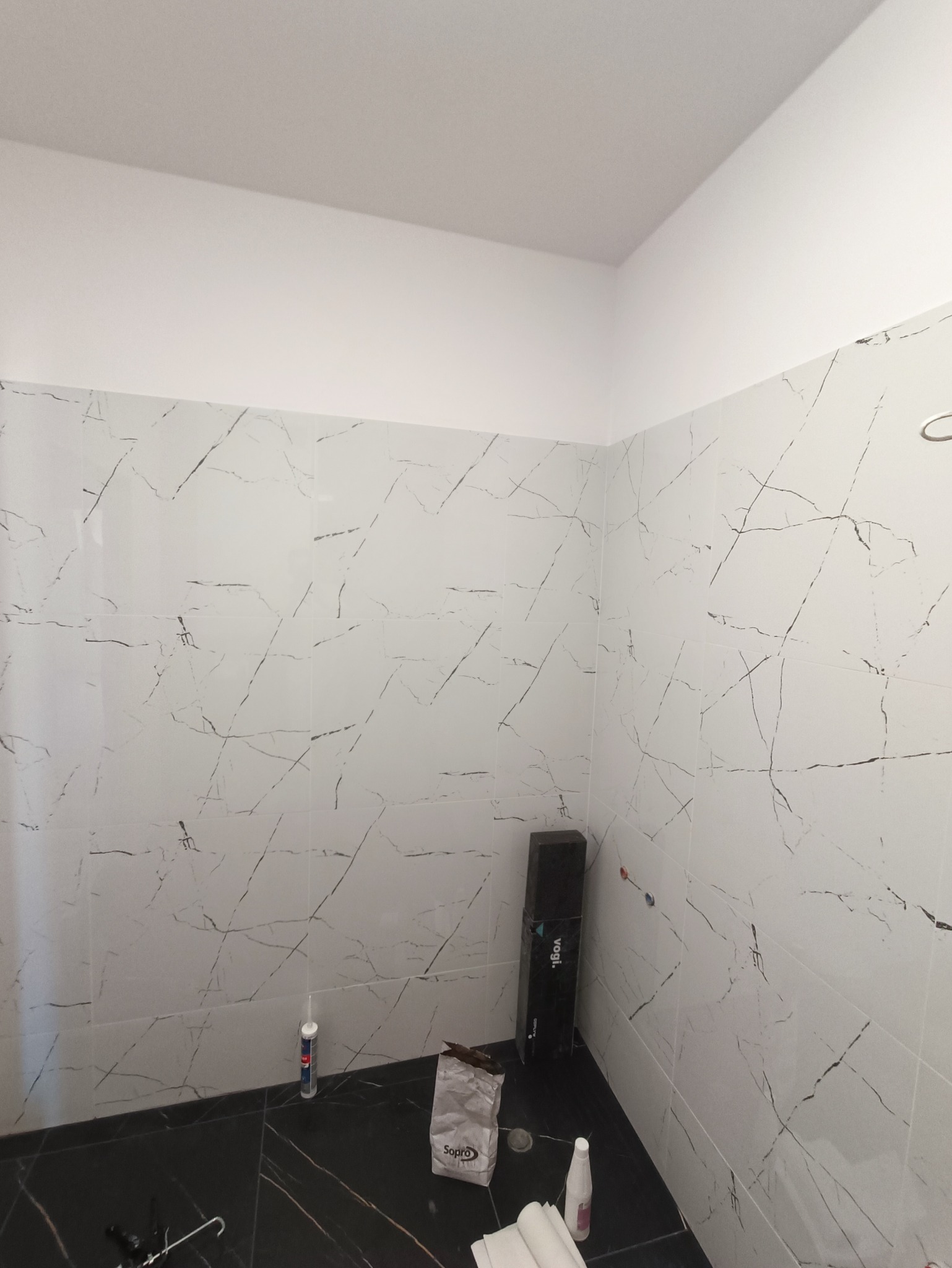 Fragment łazienki w Kotlinie z ułożonymi białymi płytkami imitującymi marmur na ścianach oraz czarnymi płytkami na podłodze, widoczne akcesoria glazurnicze i opakowanie fugi.