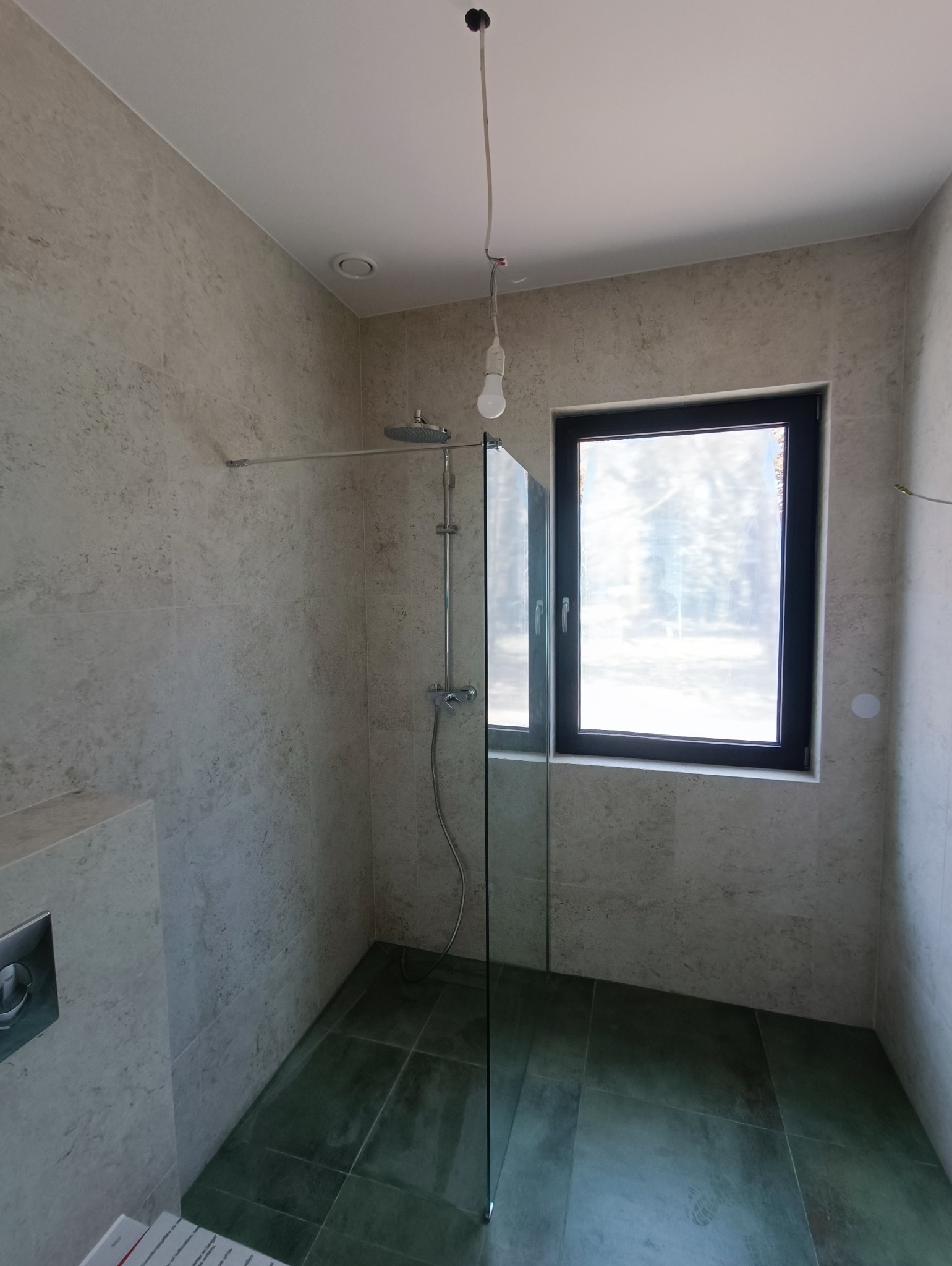 Wnętrze łazienki w trakcie wykańczania: kabina prysznicowa ze szklaną ścianką, płytki imitujące kamień na ścianach, zielone płytki na podłodze, wisząca żarówka bez klosza. Widoczne niedokończone...