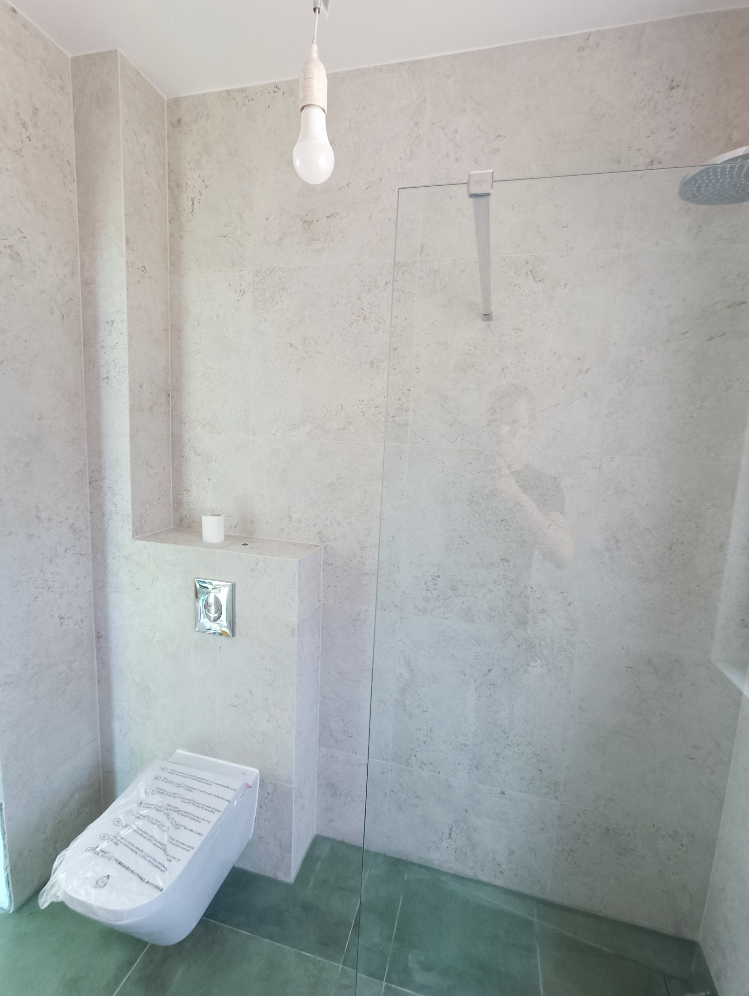 Nowoczesna łazienka z kabiną prysznicową typu walk-in, wiszącą toaletą w folii ochronnej, wykończona płytkami imitującymi kamień w odcieniach szarości i zieleni, z surową żarówką na kablu.