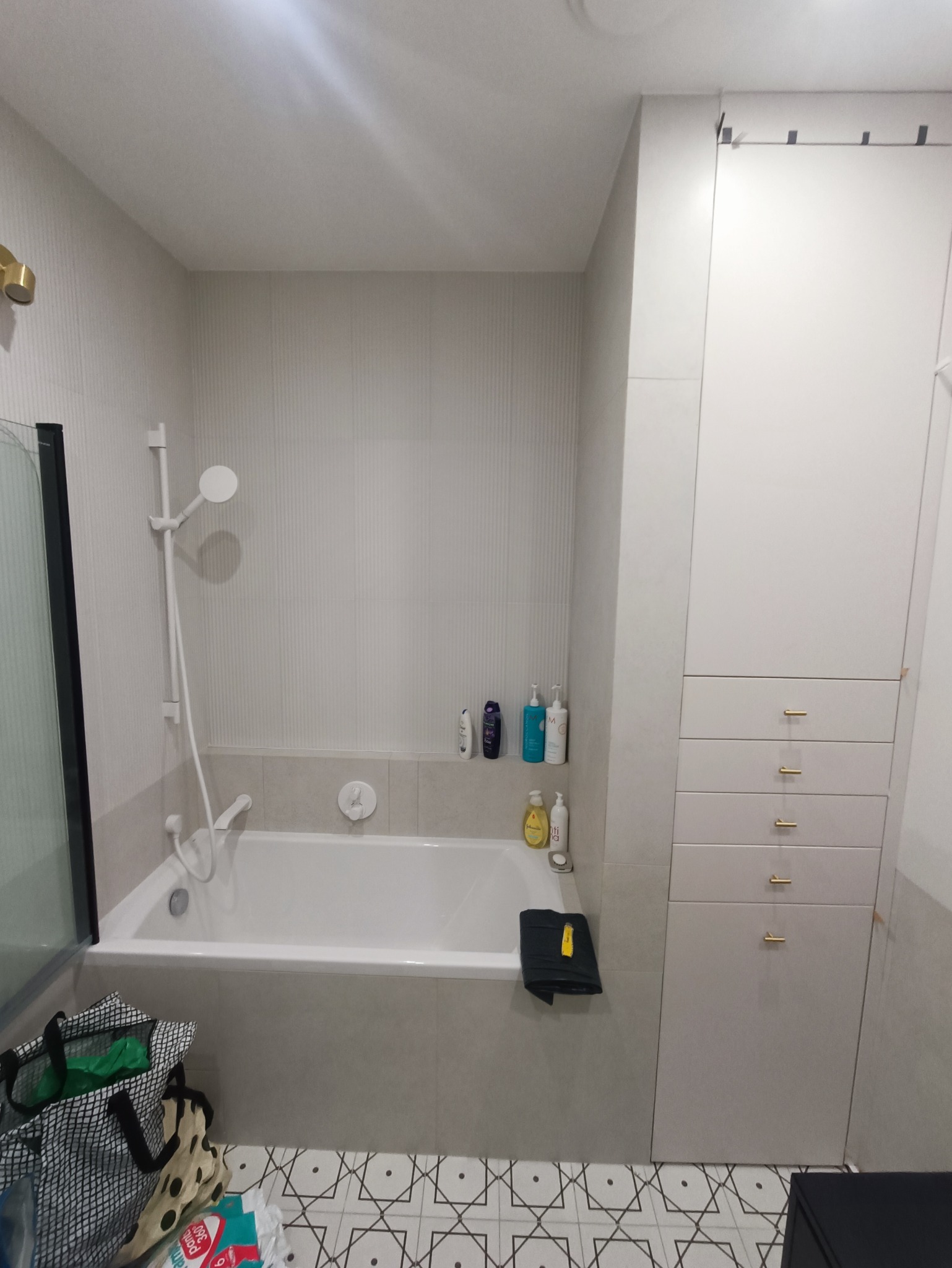 Jasna łazienka po remoncie z białą wanną, prysznicem, szafką z szufladami i geometryczną podłogą. Na wannie leży czarny ręcznik, a obok wanny stoi torba na zakupy.