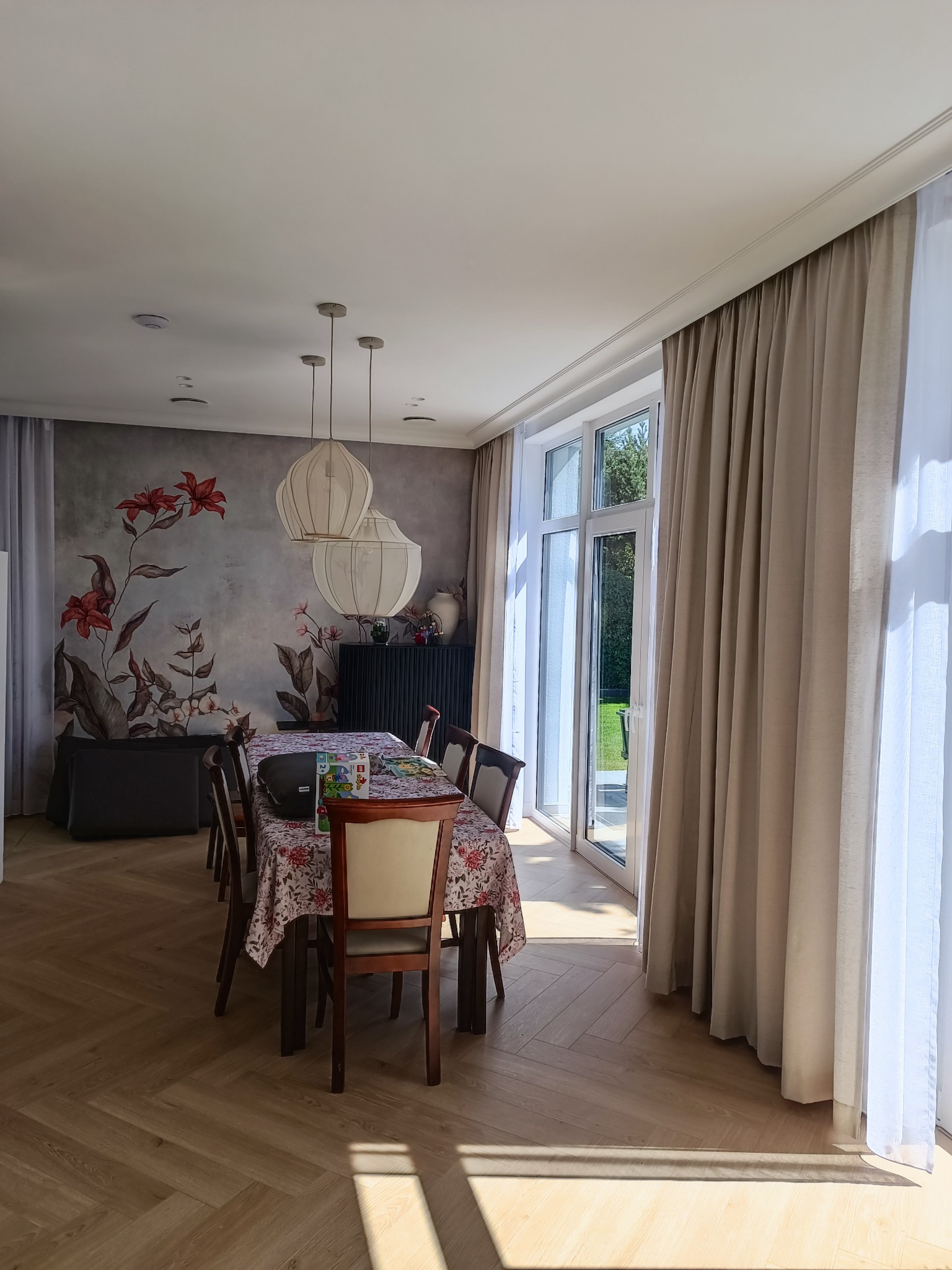 Jasne wnętrze jadalni z parkietem ułożonym w jodełkę, kwiatowym motywem na ścianie, trzema lampami wiszącymi nad stołem przykrytym kwiecistym obrusem i zasłonami w oknach.