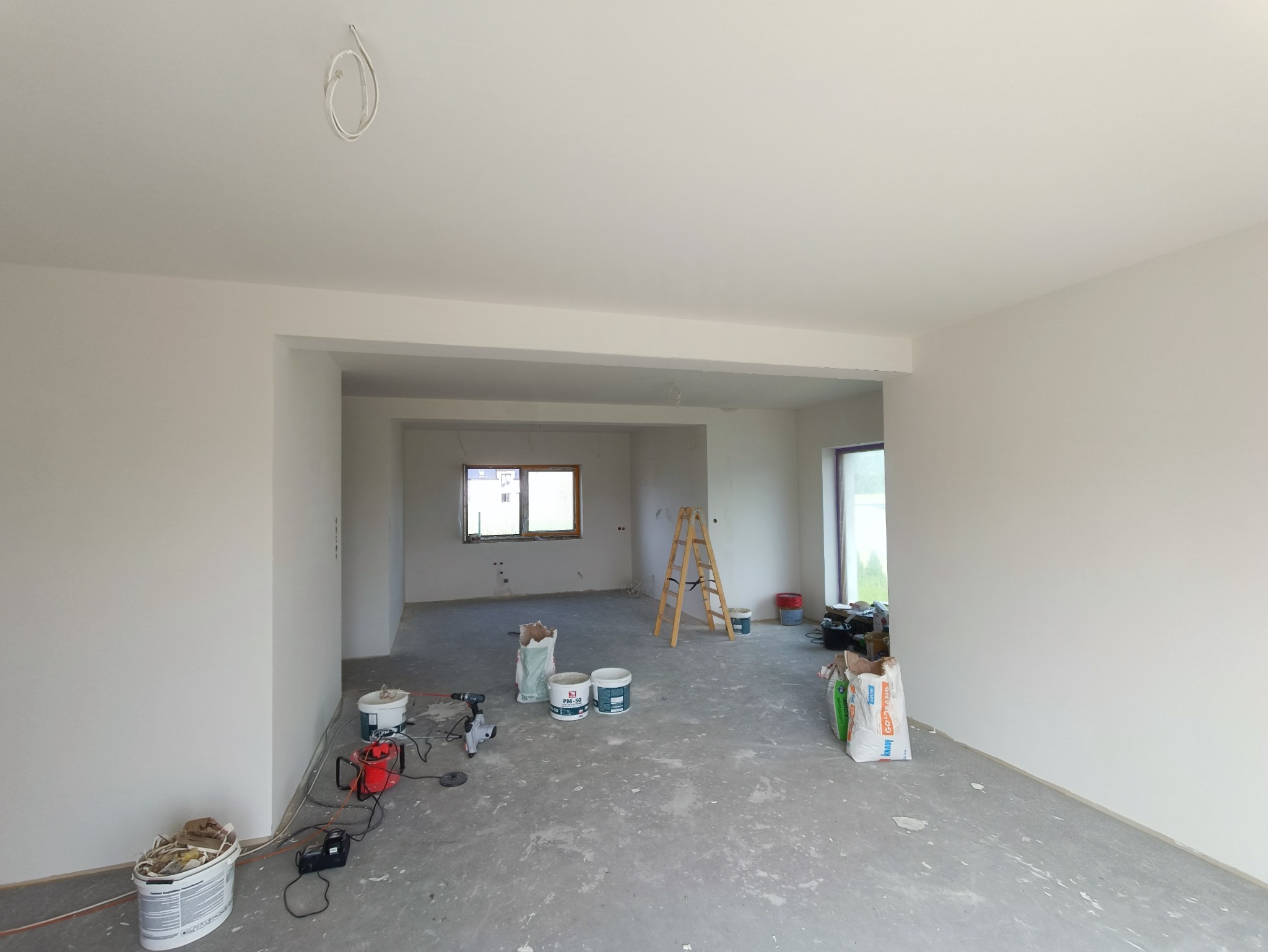 Wnętrze w trakcie prac wykończeniowych: białe ściany, drabina, worki z zaprawą, narzędzia malarskie. Widok na sąsiednie pomieszczenie z oknem.