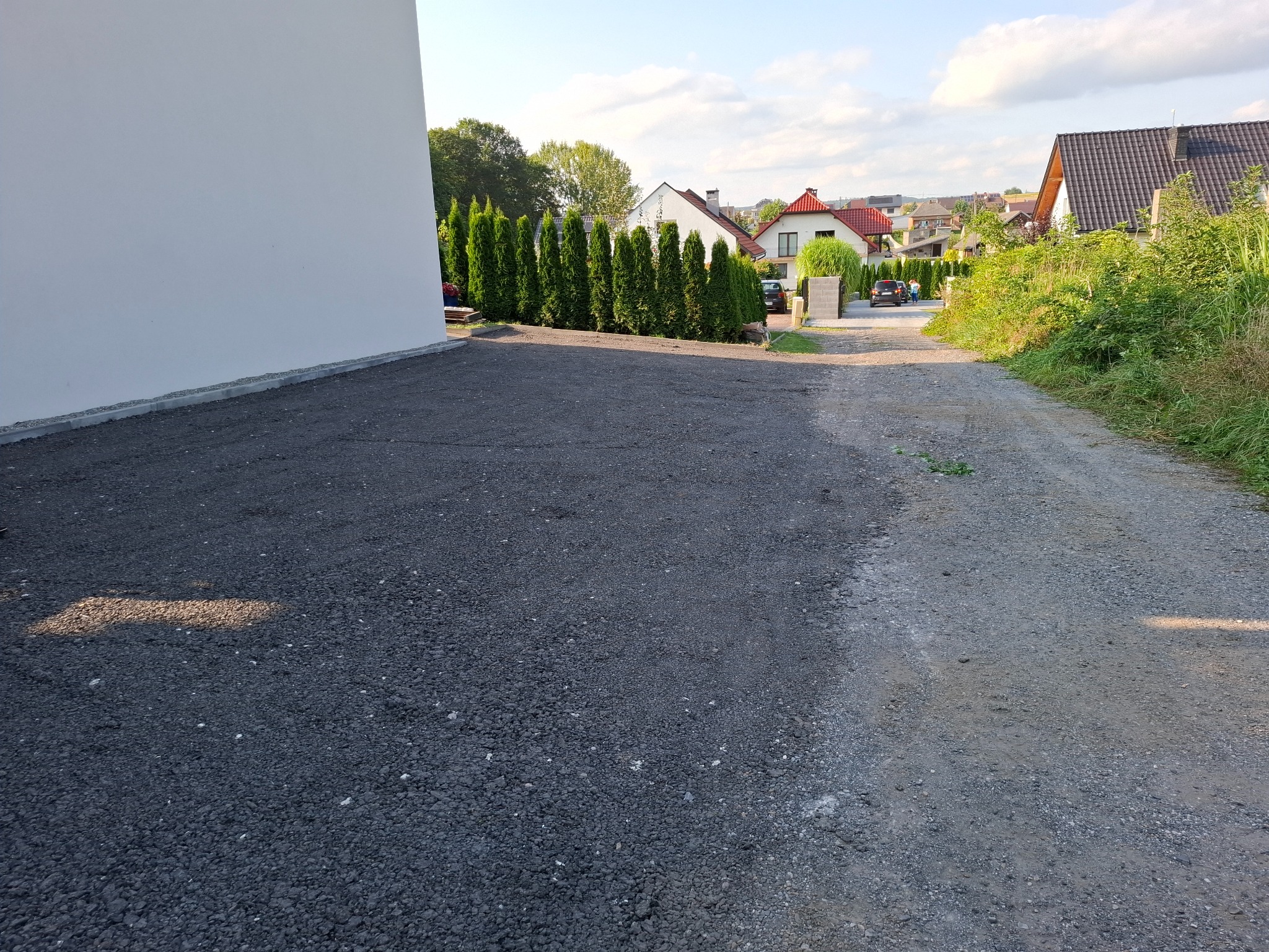 Świeżo rozsypany, ciemny tłuczeń na podjeździe przed białym budynkiem, w tle rząd tuj i domy jednorodzinne.
