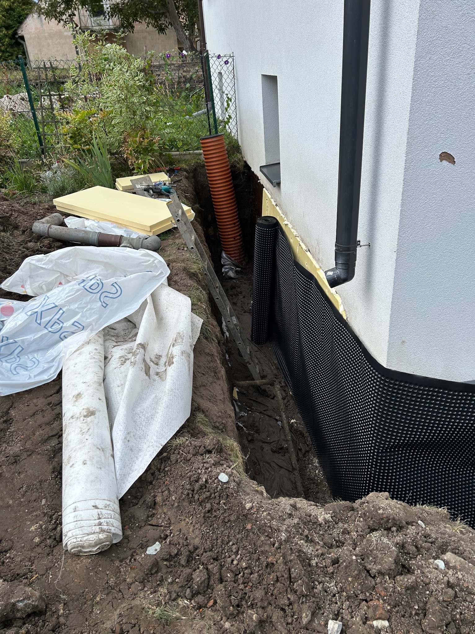 Odkopany fundament budynku z izolacją pionową z folii kubełkowej, rury drenarskie w wykopie. Widoczne materiały izolacyjne i drabina.