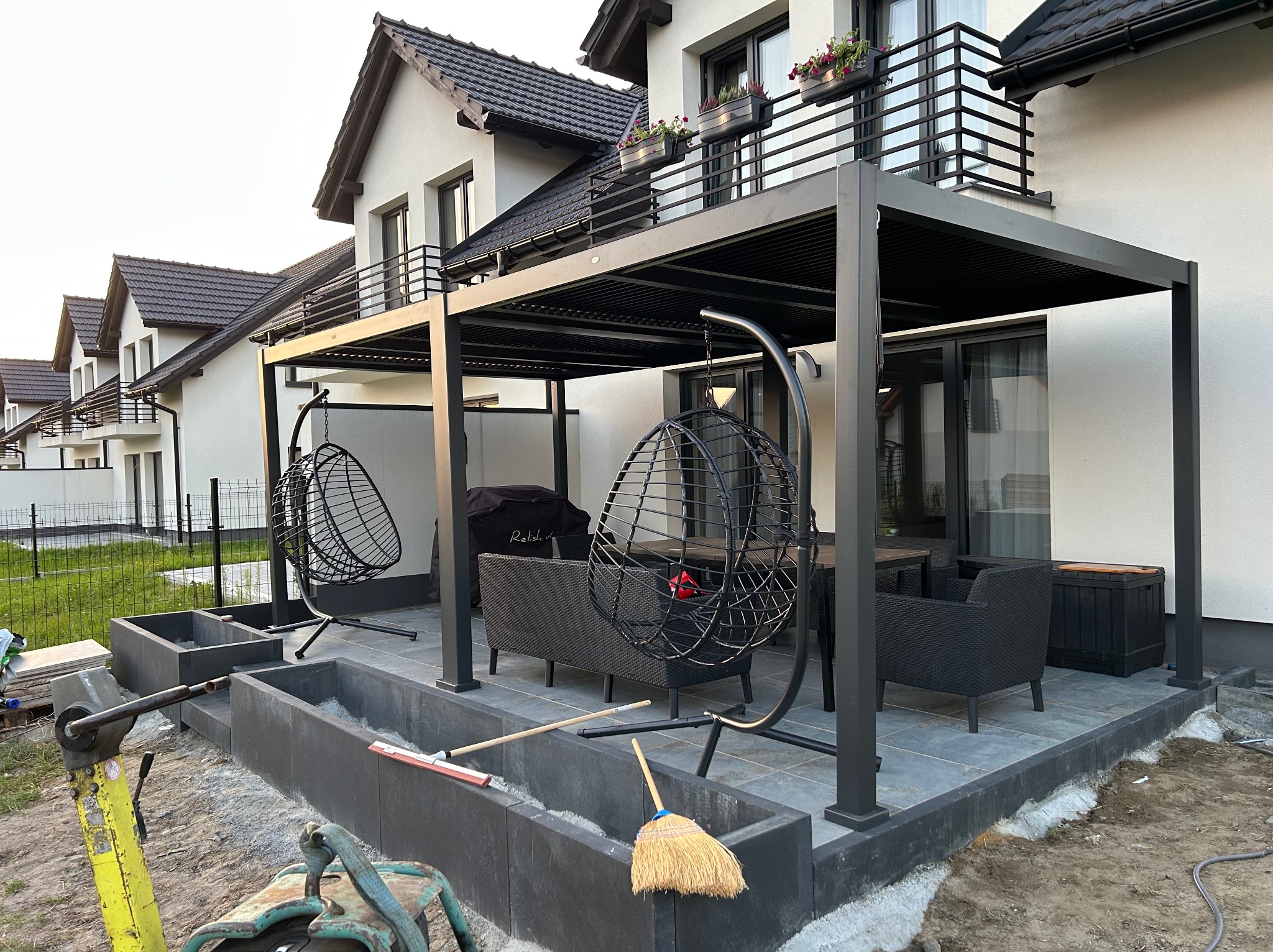 Nowoczesna pergola z czarną konstrukcją i wiszącymi fotelami, otoczona kostką brukową w trakcie układania. W tle domy jednorodzinne.