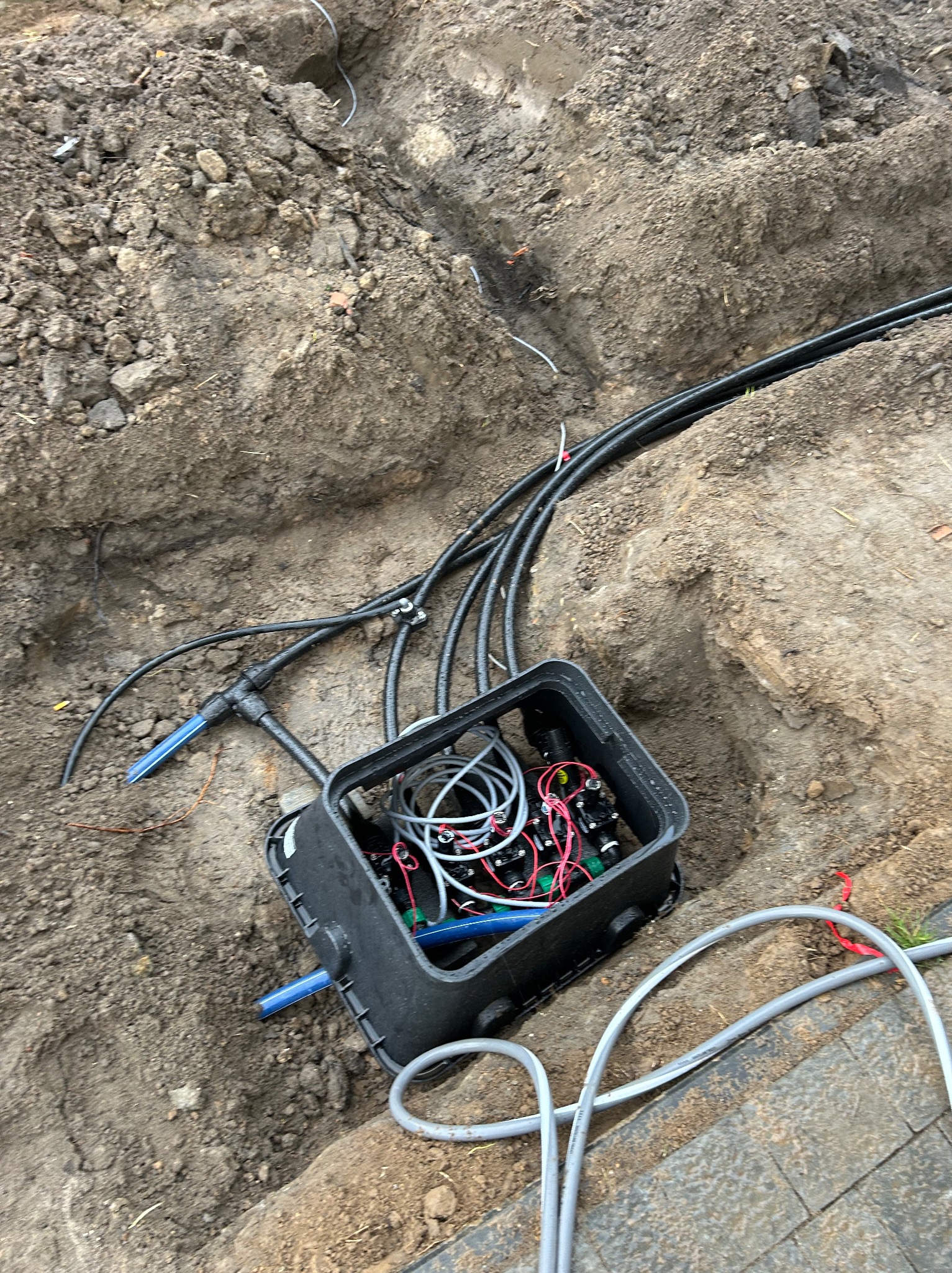 Otwarta skrzynka z instalacją elektryczną i rurami w wykopie ziemnym. Widoczne przewody, złączki i fragment chodnika z kostki.