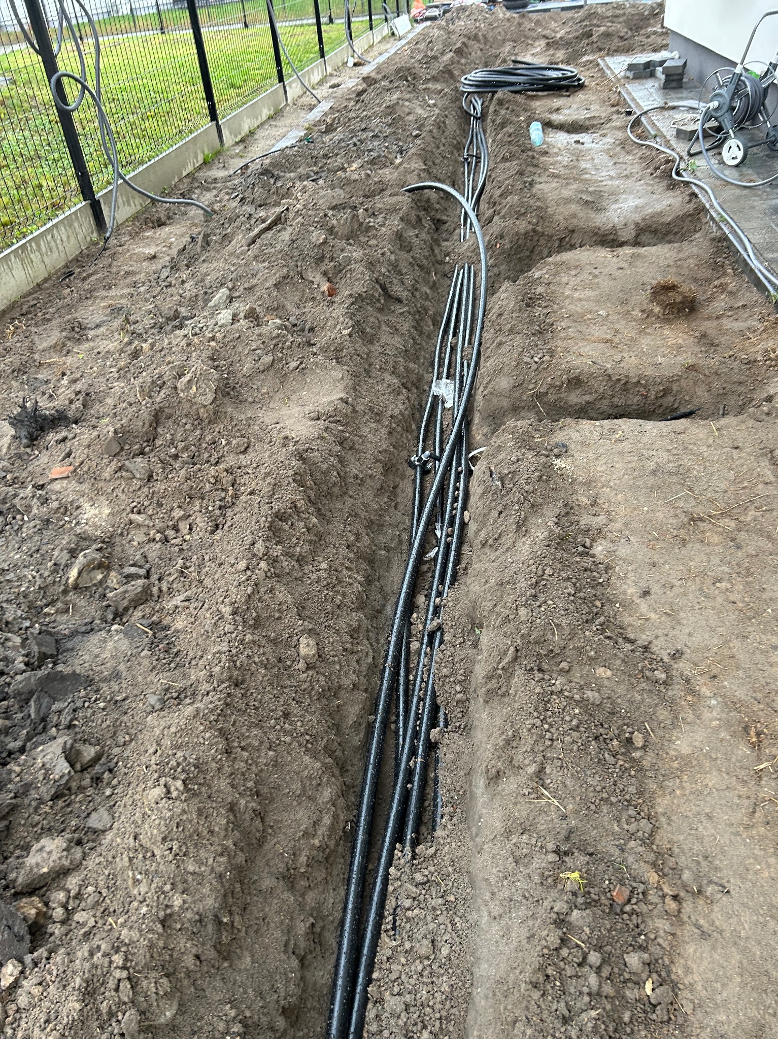 Wykop pod instalacje: czarne rury ułożone w wąskim wykopie w ziemi, częściowo zasypane, widoczne narzędzia i ogrodzenie w tle.