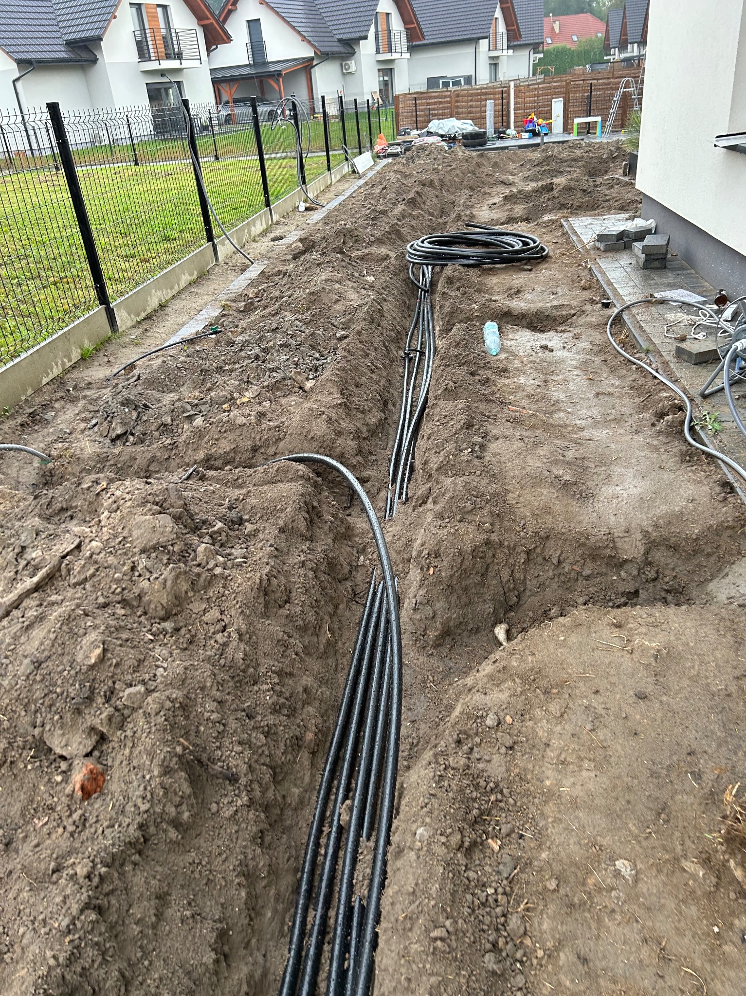 Wykop w ziemi z ułożonymi czarnymi rurami instalacyjnymi, widoczne budynki w tle i fragment ogrodzenia.
