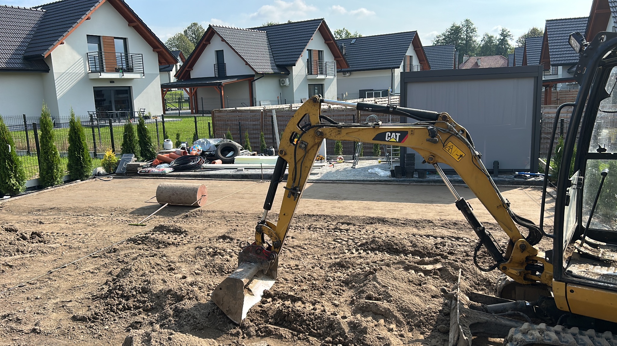 Żółta koparka CAT na placu budowy, w tle domy jednorodzinne i trawnik. Widoczny walec do ubijania ziemi i materiały budowlane.