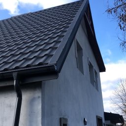 Piotr Kluka - Nowoczesny dach z blachy trapezowej w kolorze grafitowym, zamontowane rynny, surowy budynek w trakcie wykończenia elewacji, błękitne niebo w tle.
