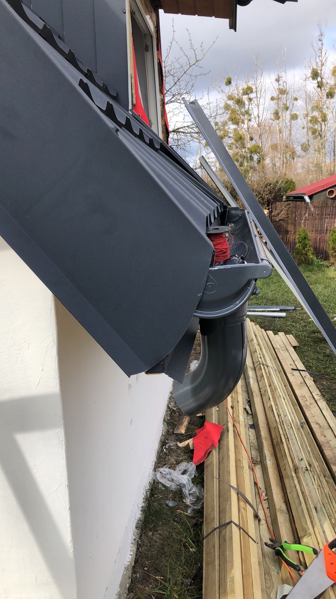 Fragment dachu z zamontowaną szarą rynną spustową, widoczny zwój czerwonego drutu w rynnie, deski i narzędzia obok ściany budynku.
