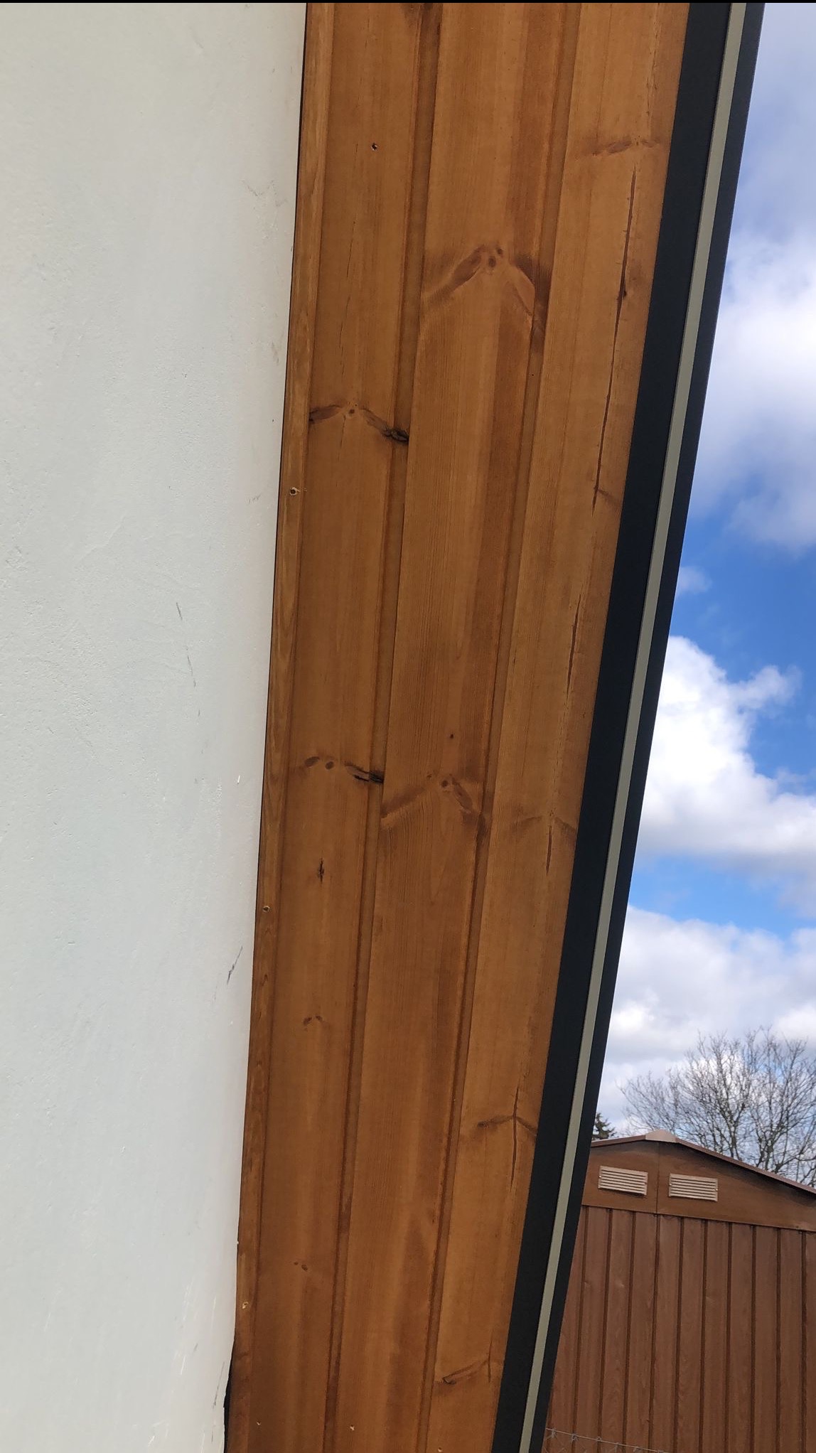Fragment dachu z drewnianą boazerią w kolorze miodowym, kontrastujący z białą ścianą i czarną listwą wykończeniową, na tle błękitnego nieba z chmurami i fragmentu brązowego budynku gospodarczego.
