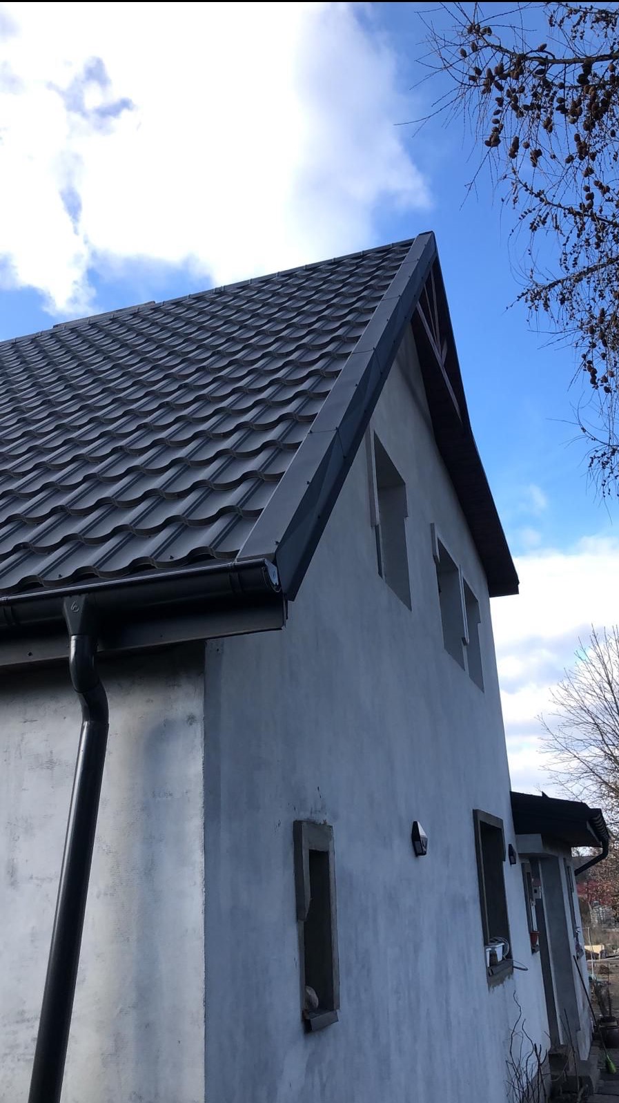 Nowoczesny dach z blachy trapezowej w kolorze grafitowym, zamontowane rynny, surowy budynek w trakcie wykończenia elewacji, błękitne niebo w tle.