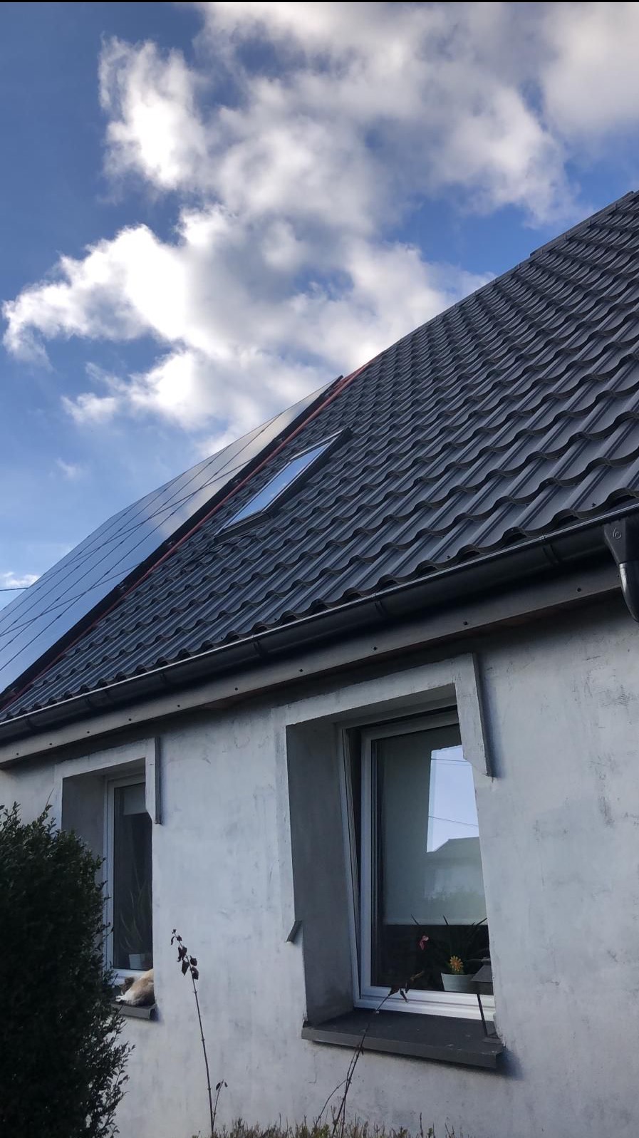 Szary dach z dachówki ceramicznej z oknem dachowym i panelami słonecznymi, widok z dołu na tle błękitnego nieba z chmurami, fragment ściany budynku z dwoma oknami.