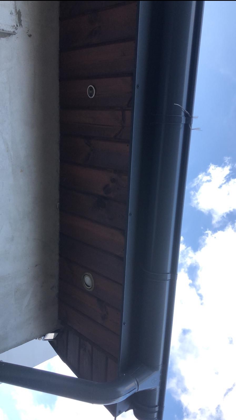 Zamontowana, grafitowa rynna na tle drewnianego okapu z wbudowanym oświetleniem punktowym, widok z dołu na błękitne niebo z chmurami.