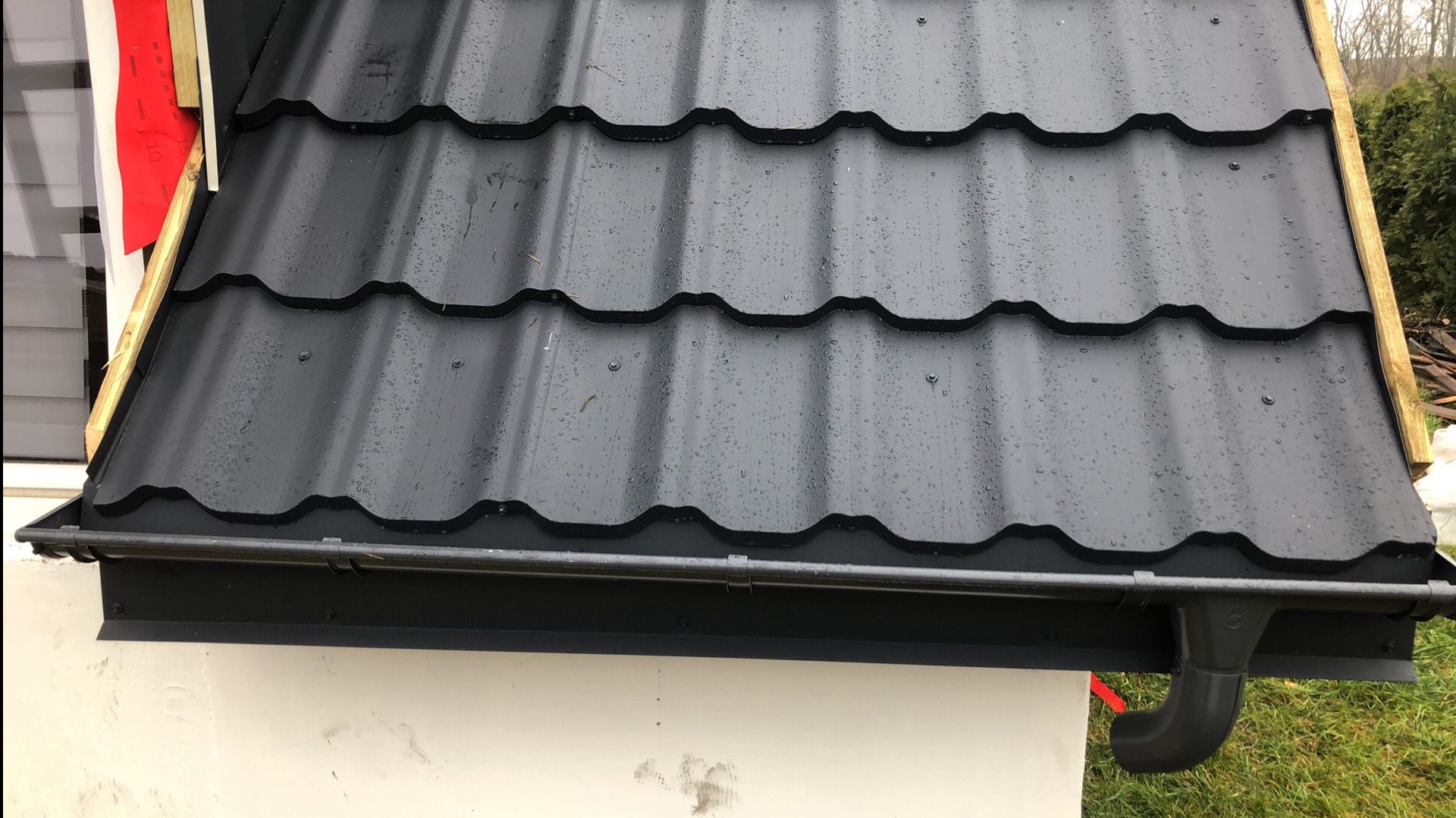 Częściowy widok dachu pokrytego czarną blachodachówką z zamontowaną czarną rynną i rurą spustową, widoczne krople deszczu na powierzchni dachu, fragment ściany budynku i elementy konstrukcji...