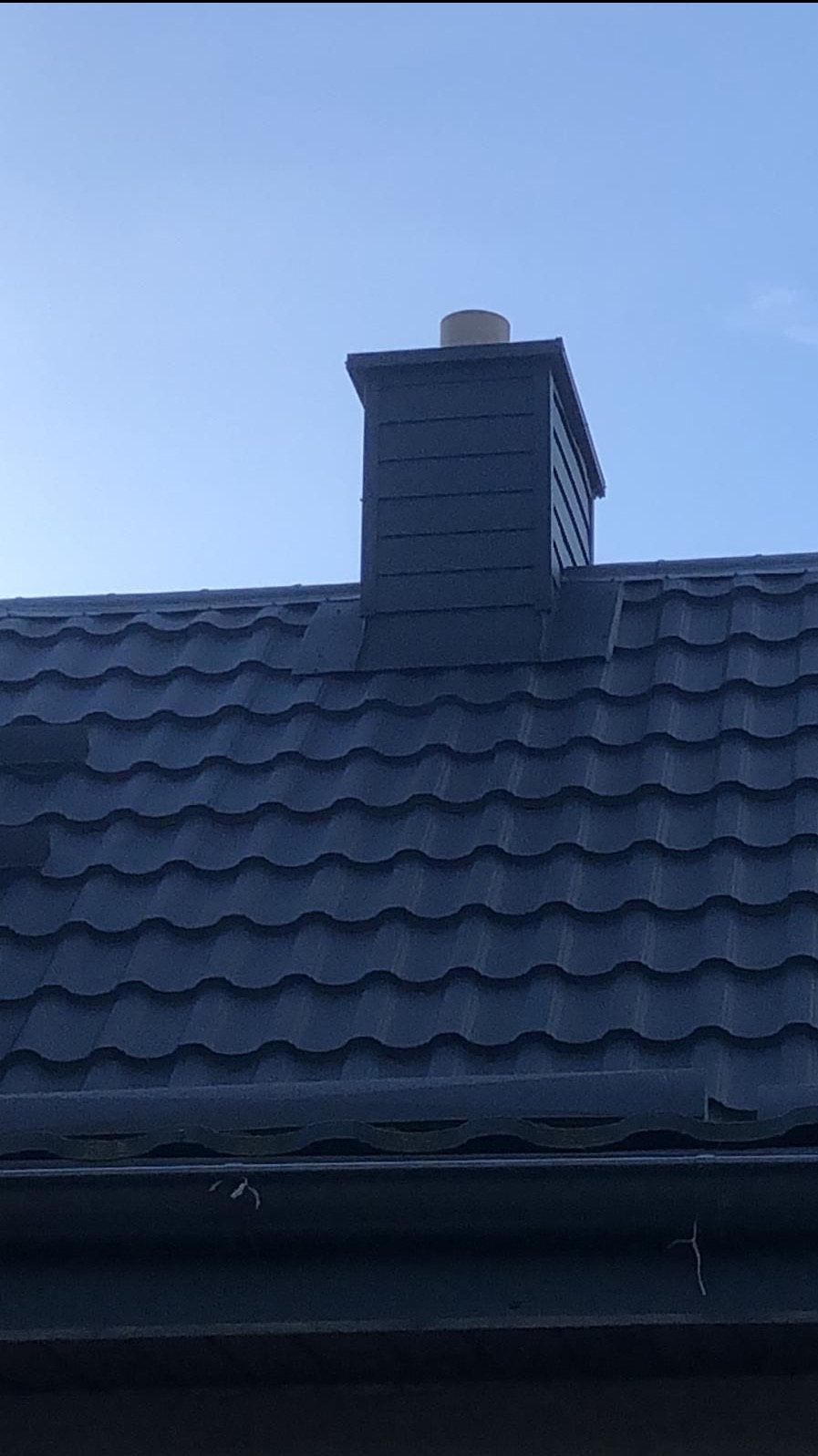 Ciemna blachodachówka z kominem w kolorze, widoczna rynna, błękitne niebo w tle.