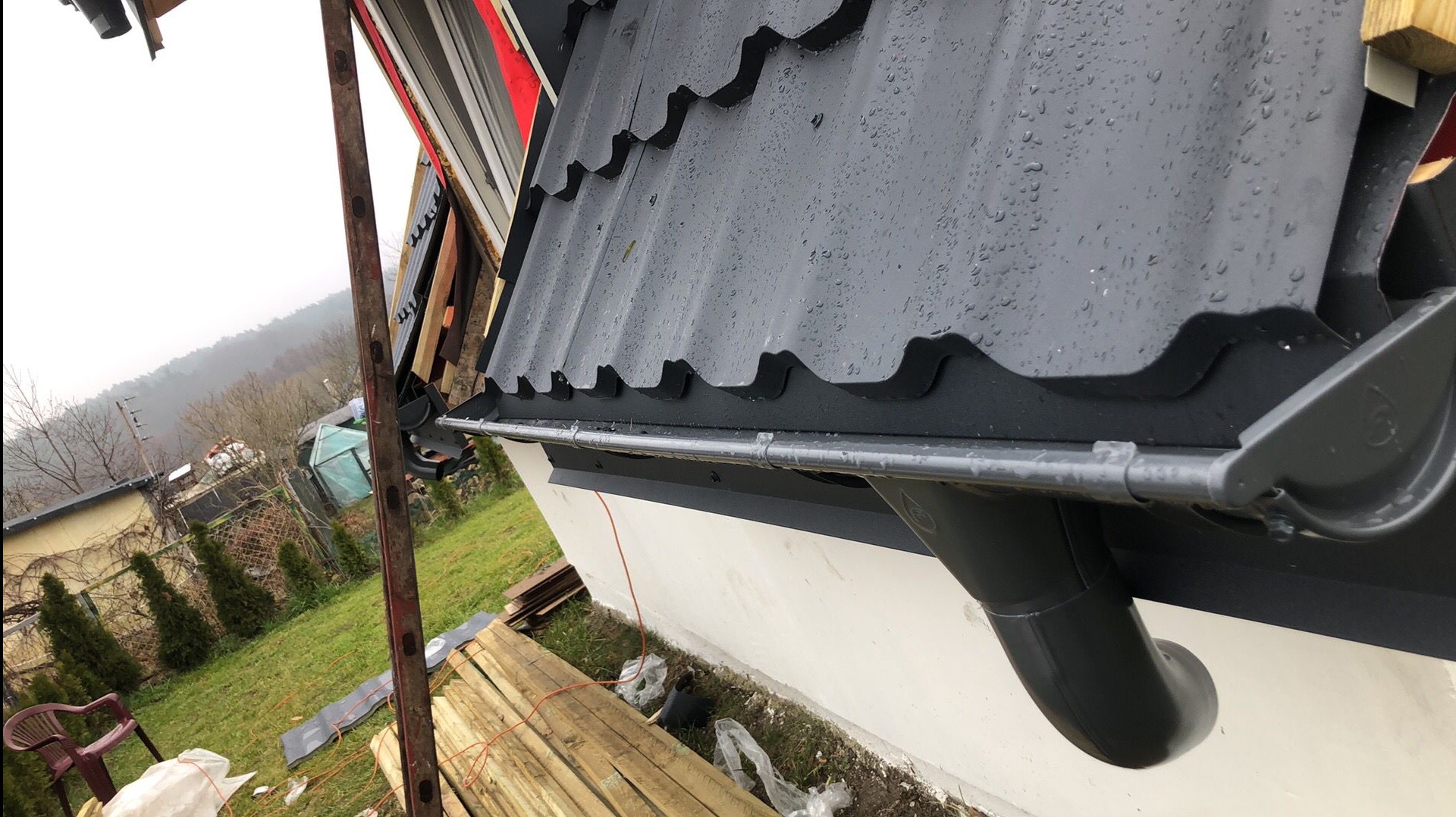 Fragment dachu pokrytego szarą blachodachówką z zamontowaną szarą rynną, na której widoczne są krople deszczu; w tle widoczny teren posesji z drzewami i budynkami.