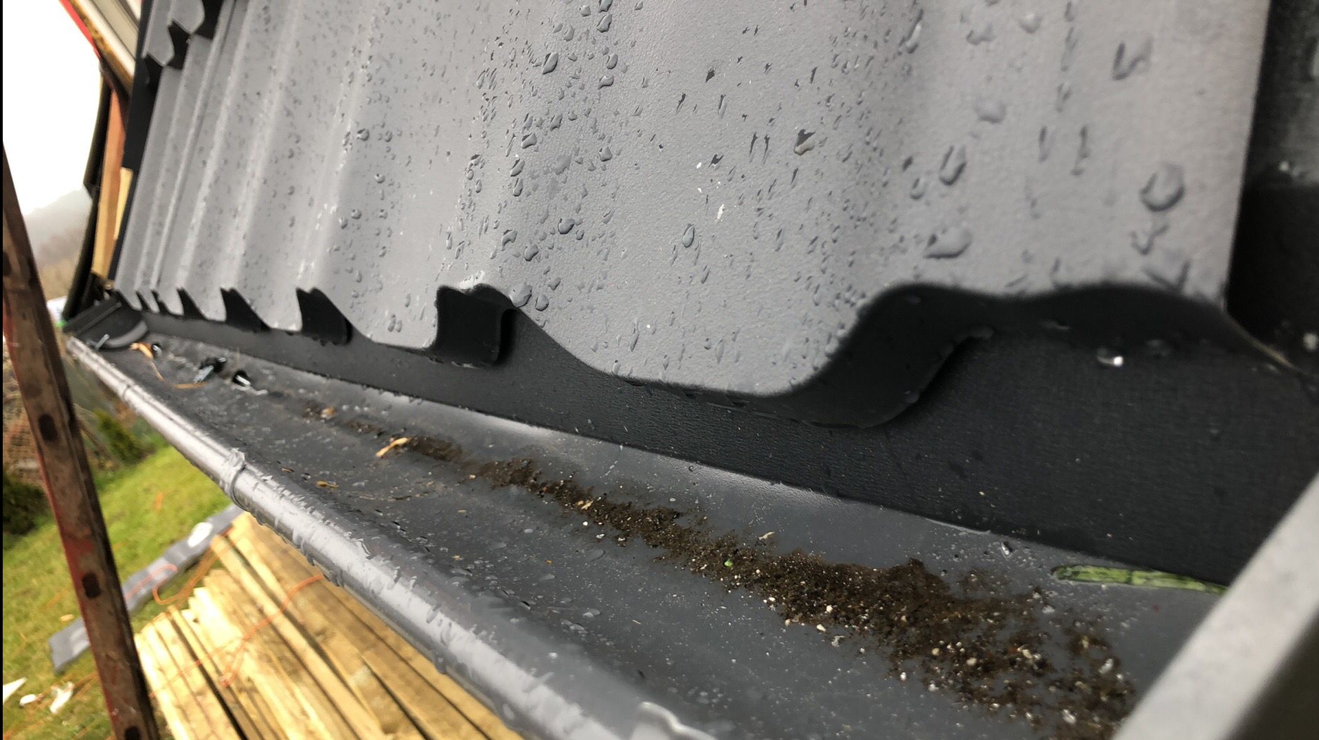 Szary dach z blachy trapezowej, widoczne krople deszczu i brudna rynna z resztkami liści i ziemi. Ujęcie z dołu, fragment rusztowania w tle.