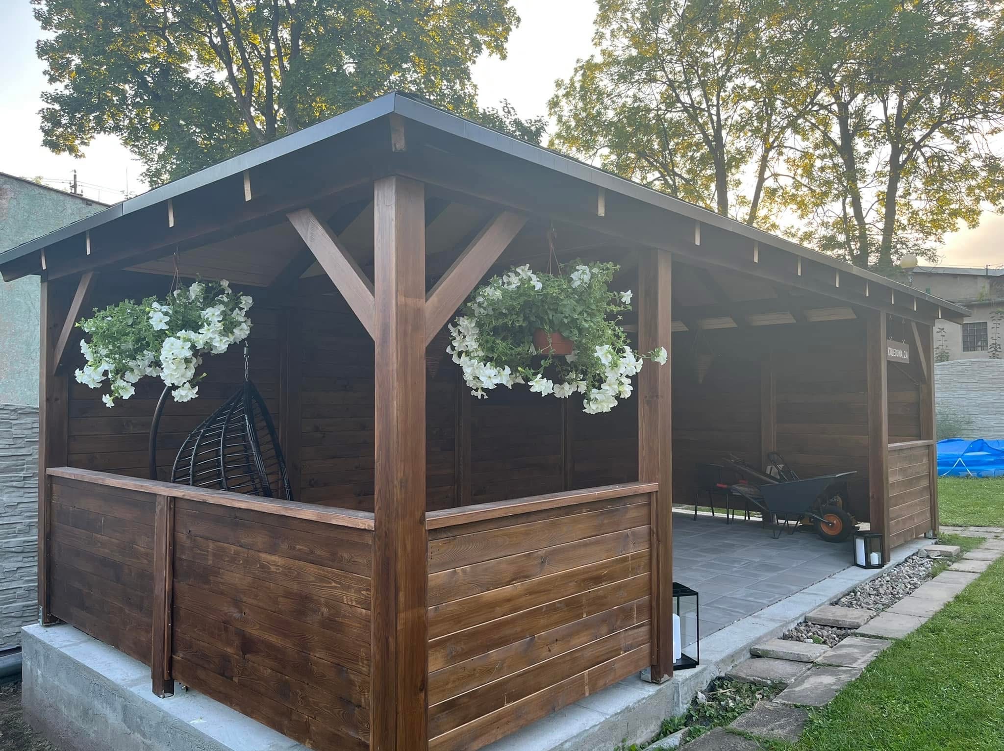 Drewniana altana z ciemnym dachem, zawieszone kwiaty i huśtawka. Na podłodze kostka brukowa, w tle zieleń i budynek. Widok na otoczenie altany.