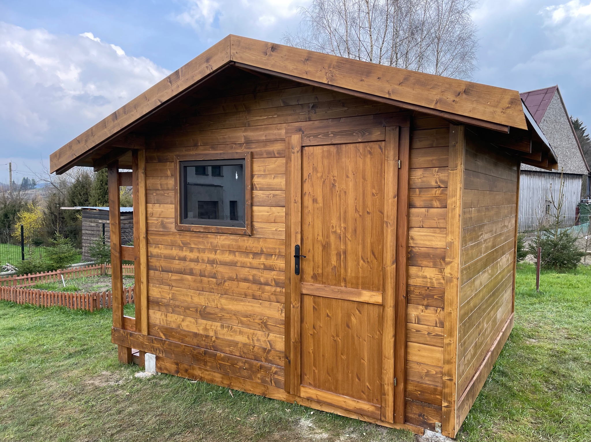 Drewniana altana ogrodowa z oknem i drzwiami, świeżo zaimpregnowana, widoczna faktura drewna. Tło: trawnik, drzewa i zabudowania.