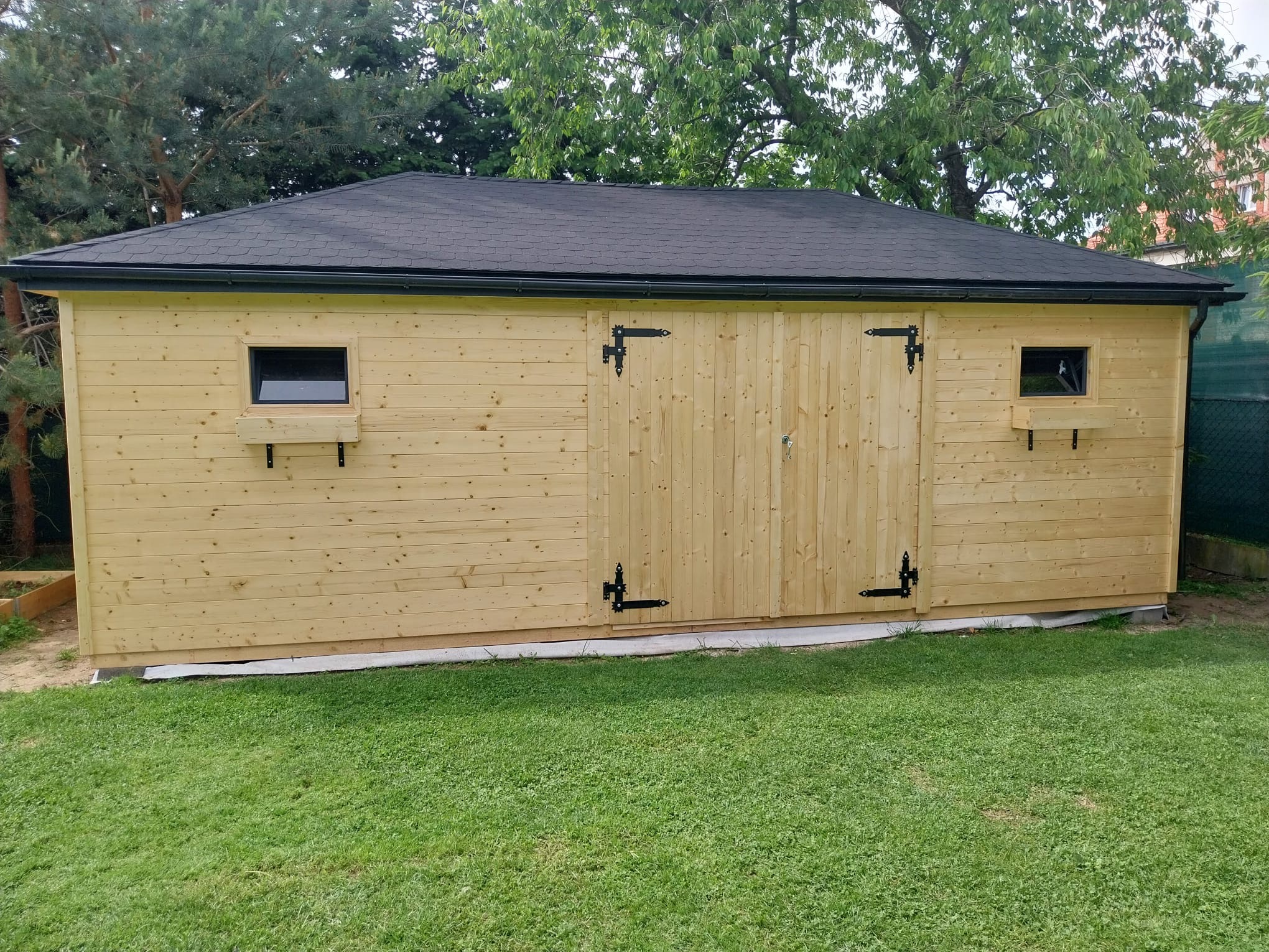 Nowoczesna, drewniana altana ogrodowa z dwuskrzydłowymi drzwiami, małymi oknami i czarnym dachem, otoczona zielonym trawnikiem i drzewami.