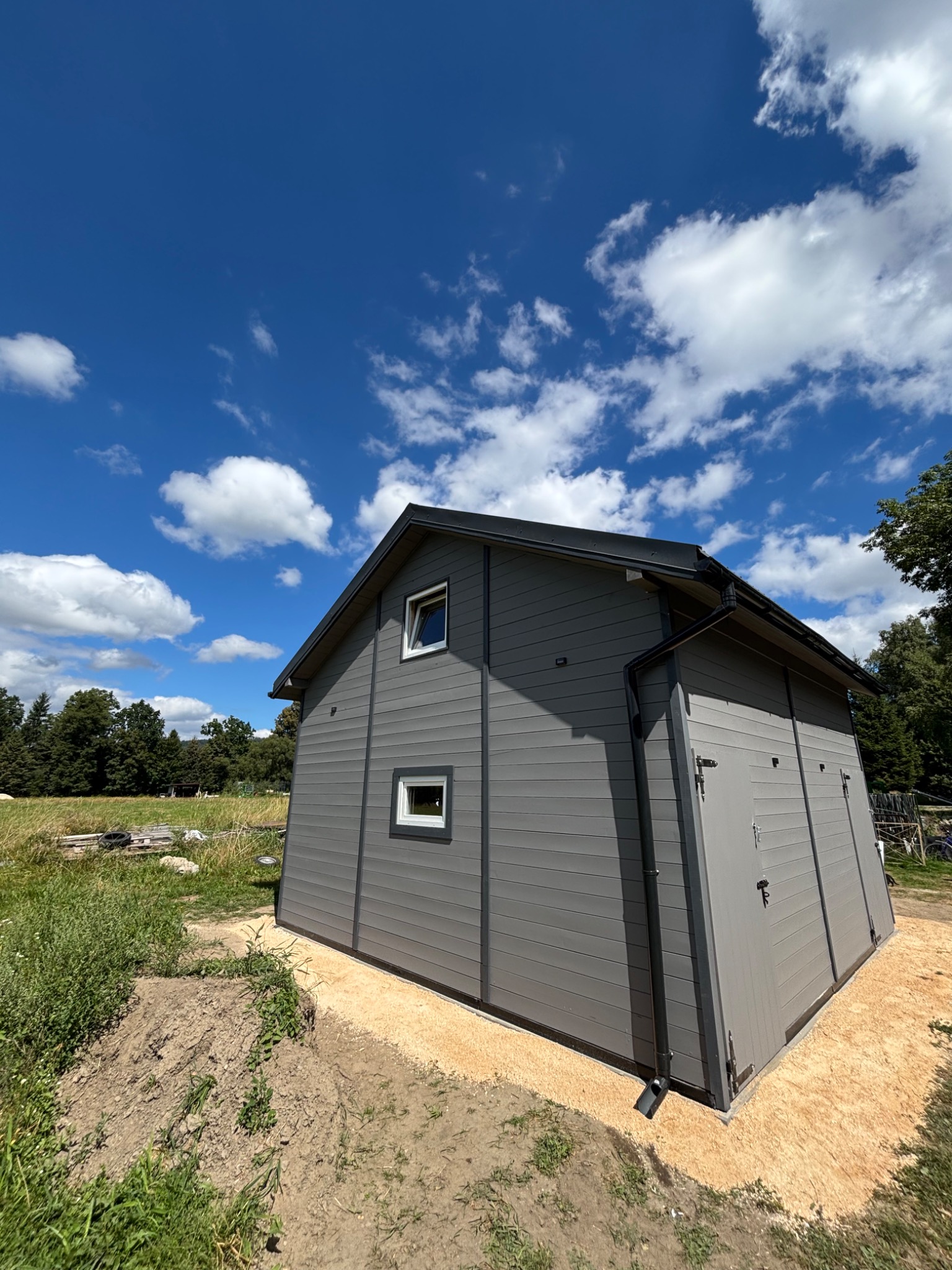Szary dom szkieletowy z widocznymi elementami wykończenia, otoczony zielenią i błękitnym niebem z chmurami. Ujęcie z dołu podkreśla bryłę budynku.