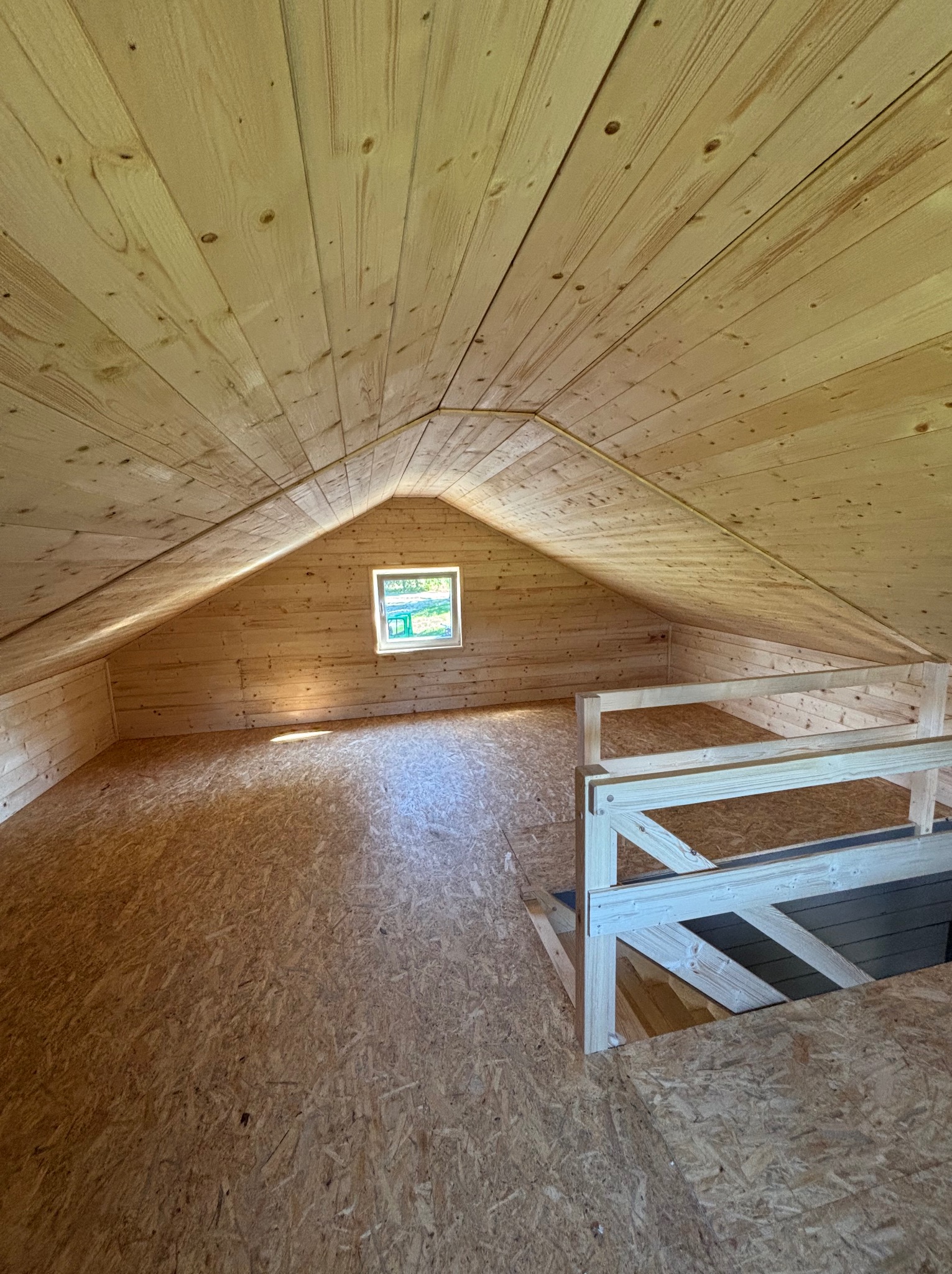 Drewniane poddasze z małym oknem i drewnianą barierką. Widoczne deski na ścianach, suficie i podłodze. Ujęcie z dołu, podkreślające przestrzeń i strukturę drewna.