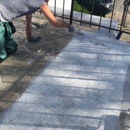 Remonty.GZ - Pracownik w szarych rękawicach nakłada żywicę na włókno szklane na balkonie, widoczne nożyczki, wałek malarski i zielone spodnie robocze.