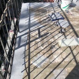 Remonty.GZ - Balkon w trakcie malowania: widoczne narzędzia malarskie, wiadro z farbą i częściowo pomalowana powierzchnia podłogi z widocznymi smugami i nierównościami, rzucane cienie krat balustrady.