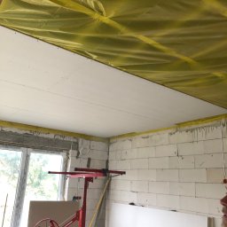 Remonty.GZ - Montaż płyty gipsowo-kartonowej na suficie, widoczna podpora montażowa, folia ochronna na stropie, surowy stan ścian z betonu komórkowego, okno z widokiem na zieleń.